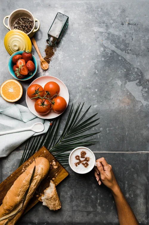 Flat lay of food items for a cookbook cover displayed on a concrete surface, including strawberries, tomatoes, an orange, bread, a cappuccino with a cinnamon Visit Tampa Bay logo design, and various bowls and utensils.
