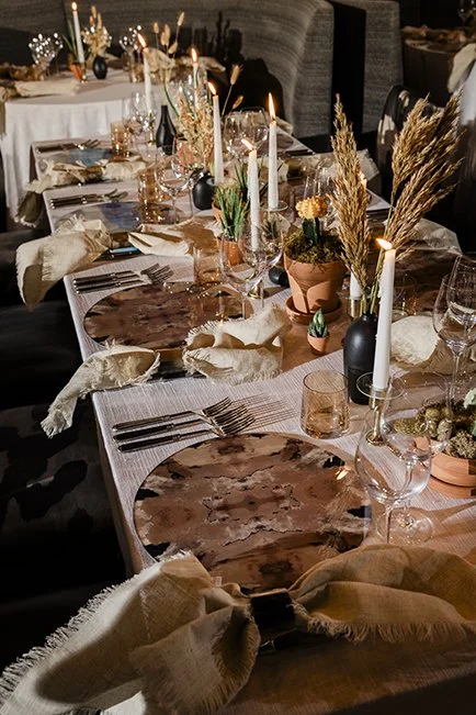 A long dining table set for an event, decorated with candles, potted plants, dried pampas grasses, and place settings including plates, glassware, and napkins in a warmly lit setting.