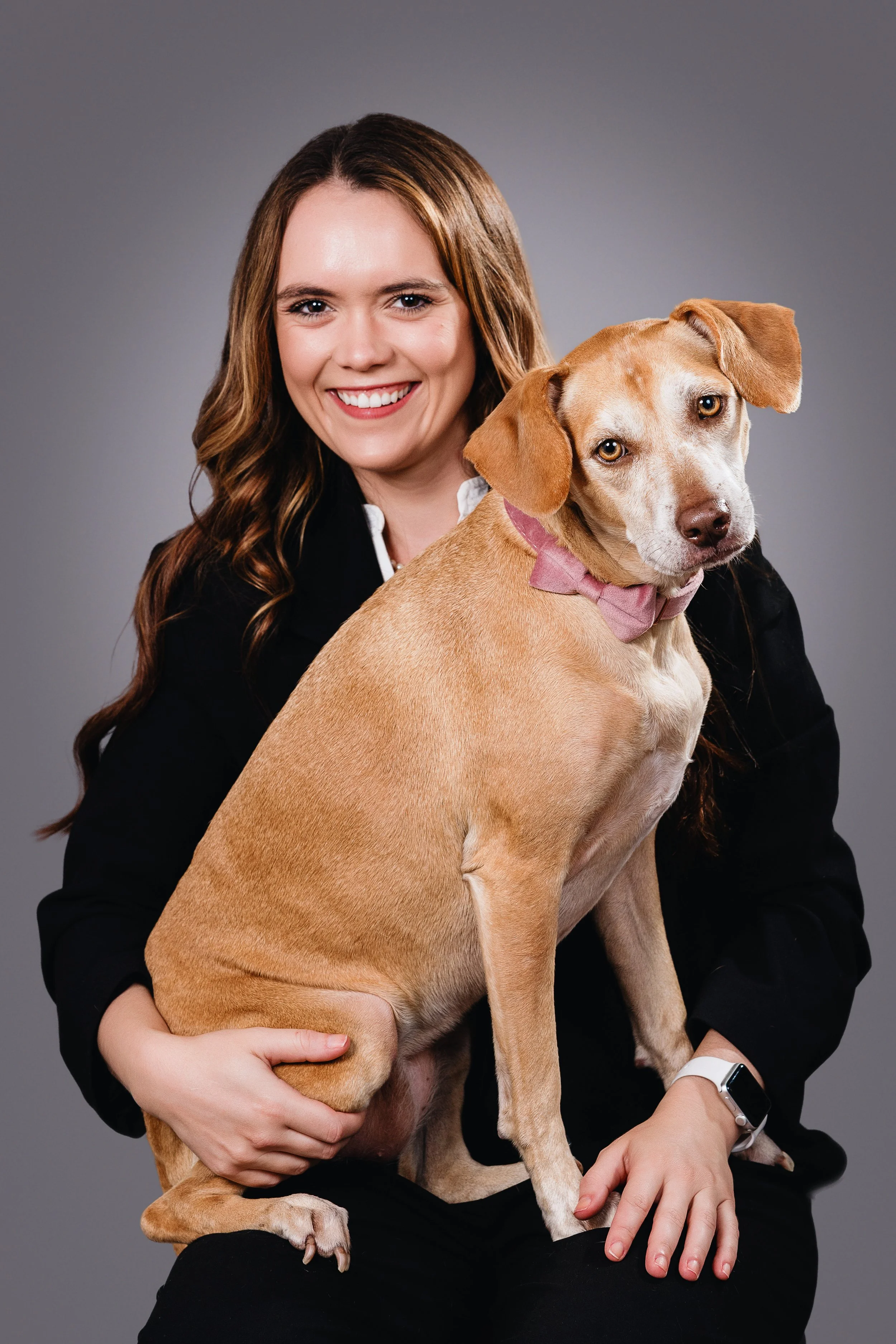 A woman with long wavy brown hair smiling, holding a tan-colored dog with a pink bow tie, both sitting against a plain gray background.