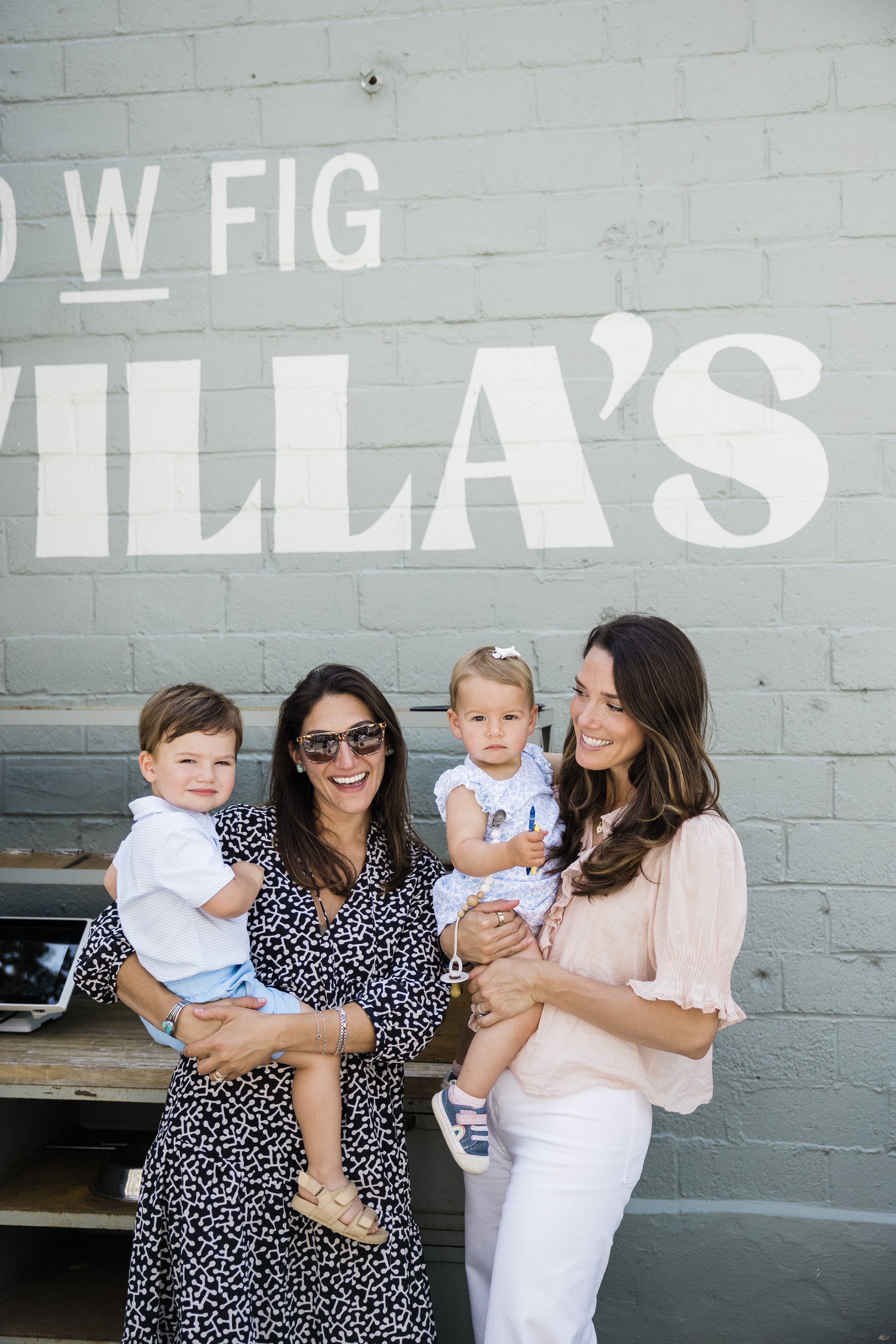 Group of three women and two children standing outside in front of a gray brick wall with white painted text that reads 'W. FIG WILLA'S.' One woman is holding a young girl, and another woman is holding a boy, all smiling and posing for the photo.