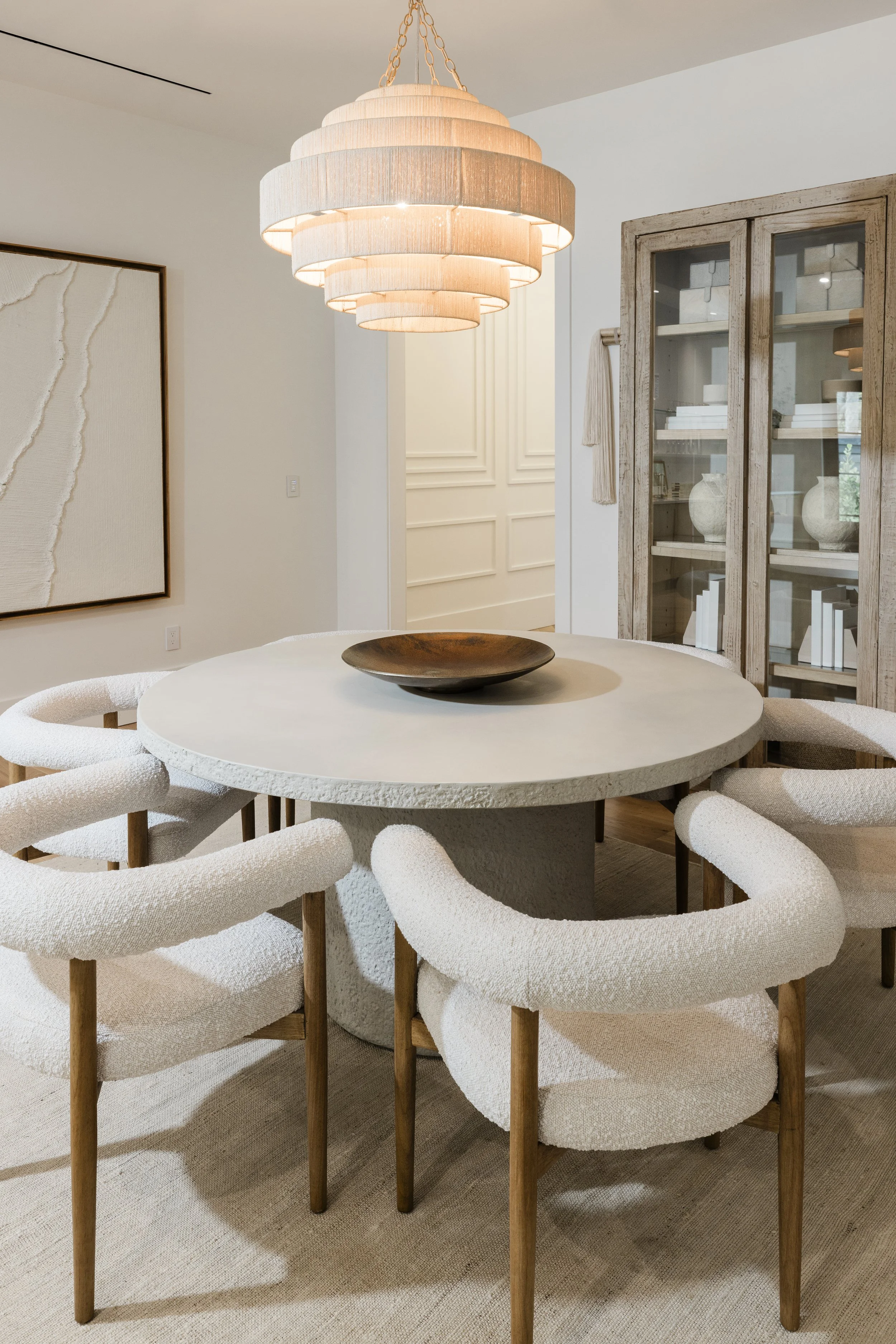 Modern dining room with a round, textured white table, surrounded by white upholstered chairs with wooden legs. A large, layered beige pendant light hangs above the table. A glass-fronted wooden cabinet displays white vases and books. The walls are w