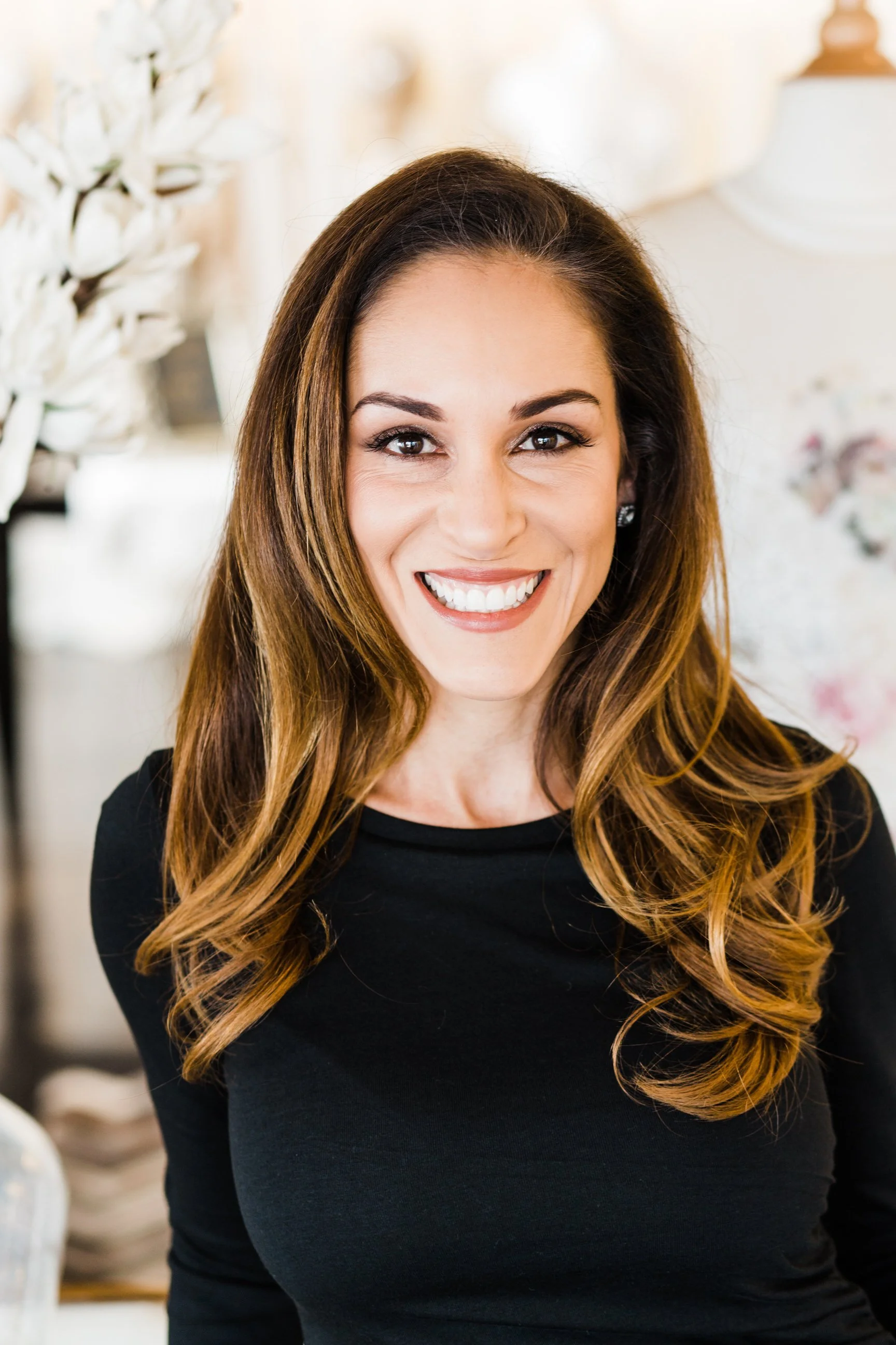 Close-up of a woman with long wavy brown hair smiling at the camera, wearing earrings and a black top, in a bright indoor setting with blurred white decor in the background.