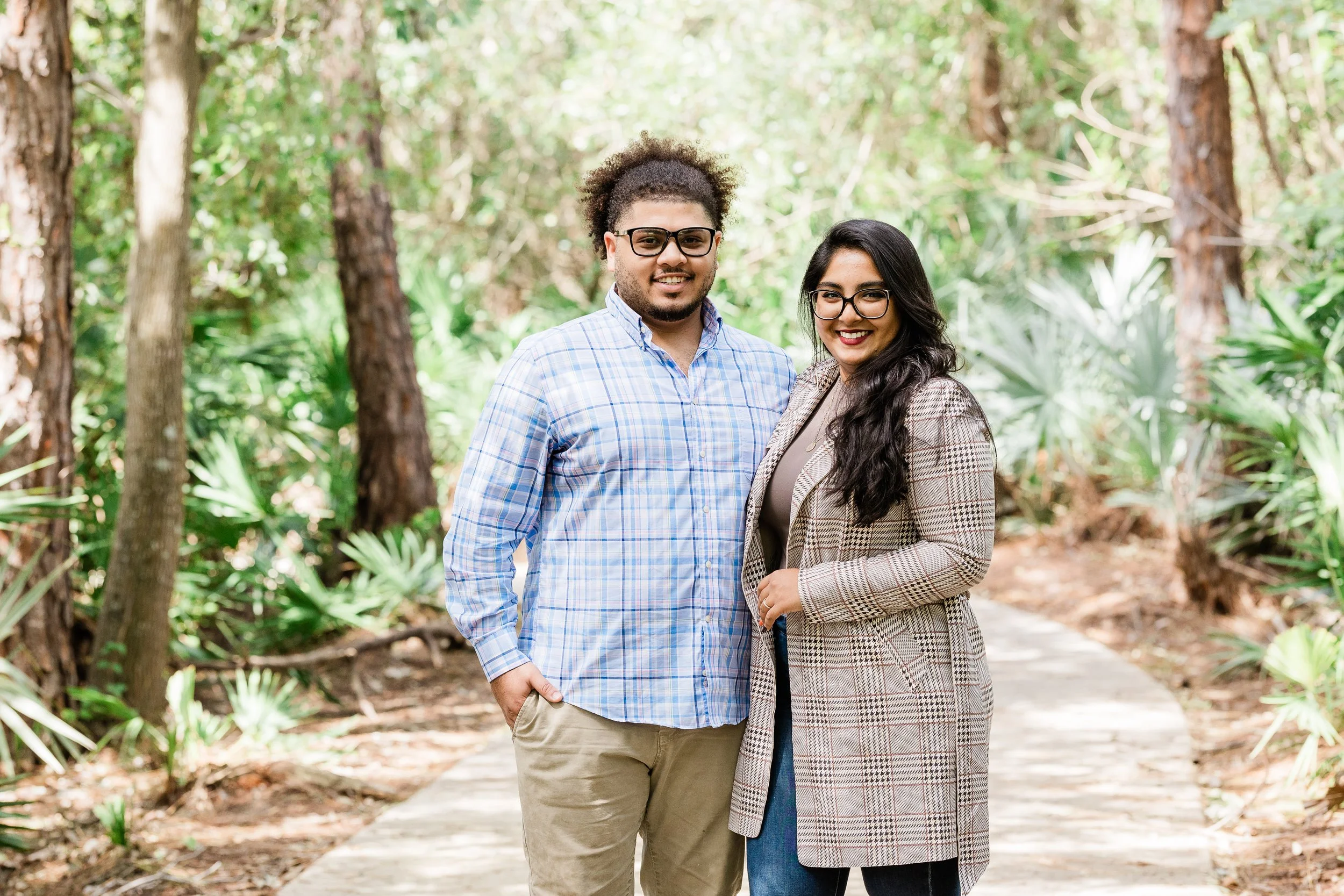 A man and woman standing together on a forest trail, smiling at the camera, surrounded by trees and green foliage.