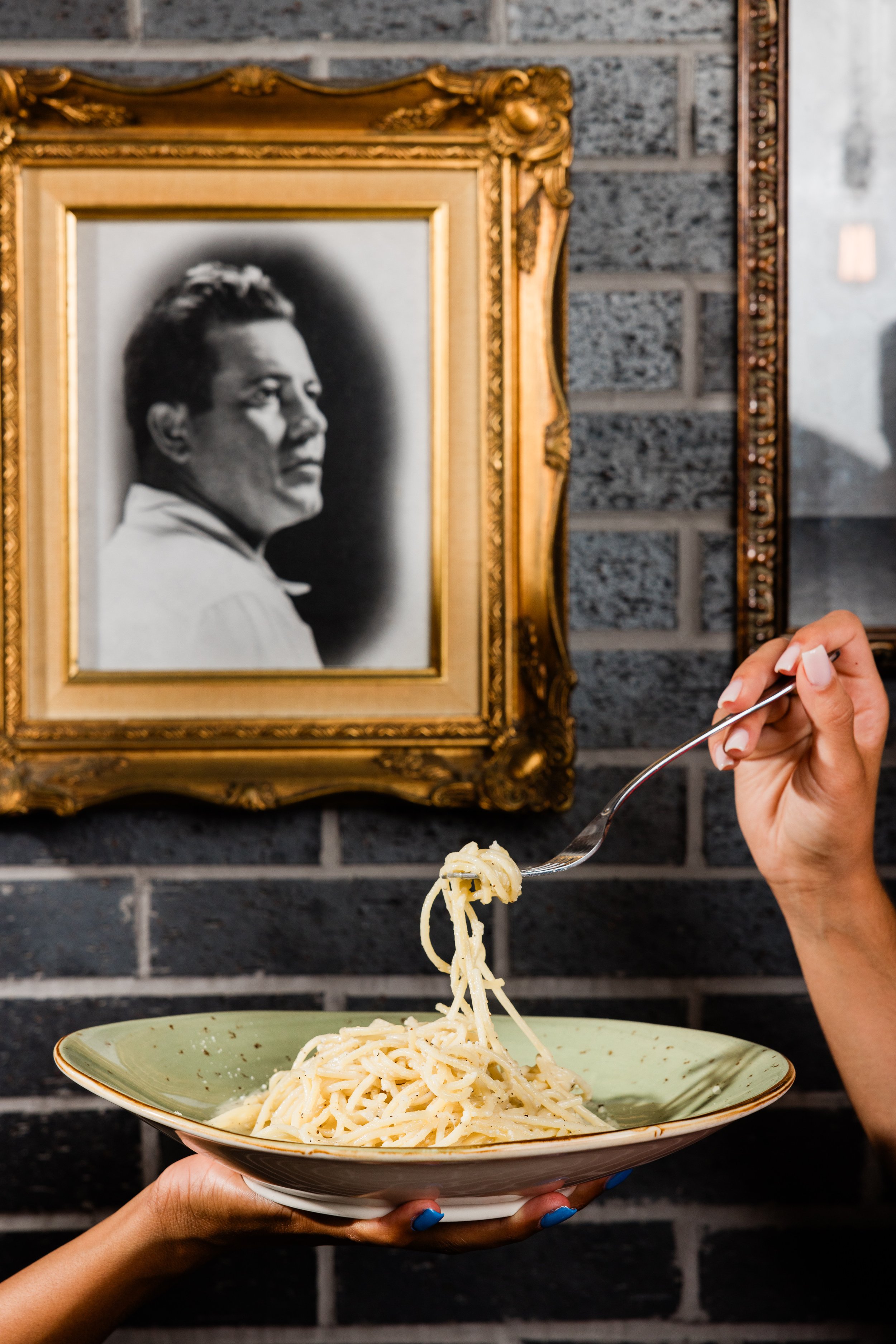 A person holding a bowl of spaghetti over a dark table with a framed black and white portrait of a man on a brick wall in the background. A hand is using a fork to lift a portion of pasta.