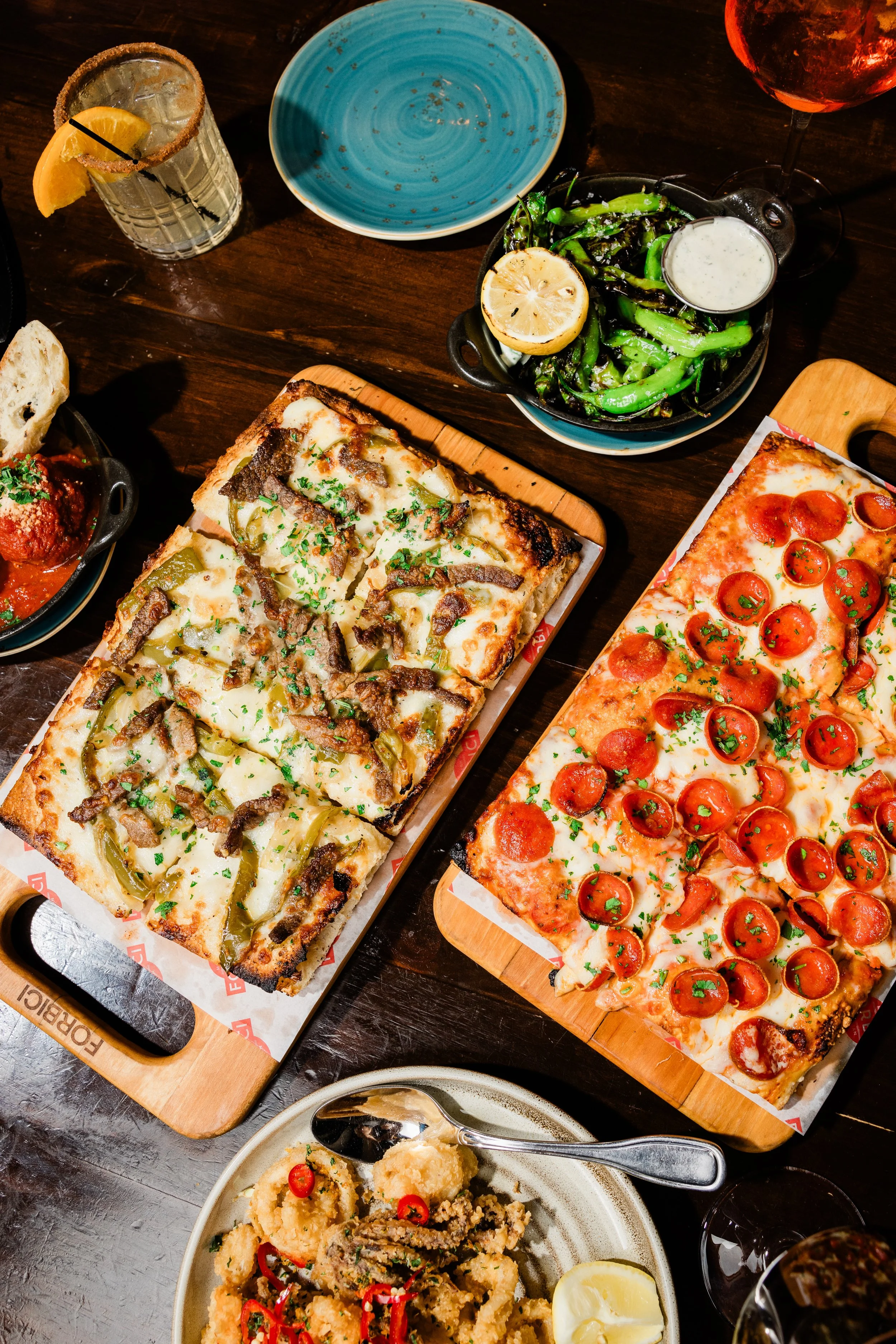 A top-down view of a dining table with various dishes including two large rectangular pizzas, a bowl of fried calamari garnished with red chili slices and lemon wedge, a plate of roasted vegetables with lemon and a cup of creamy dipping sauce, a glas