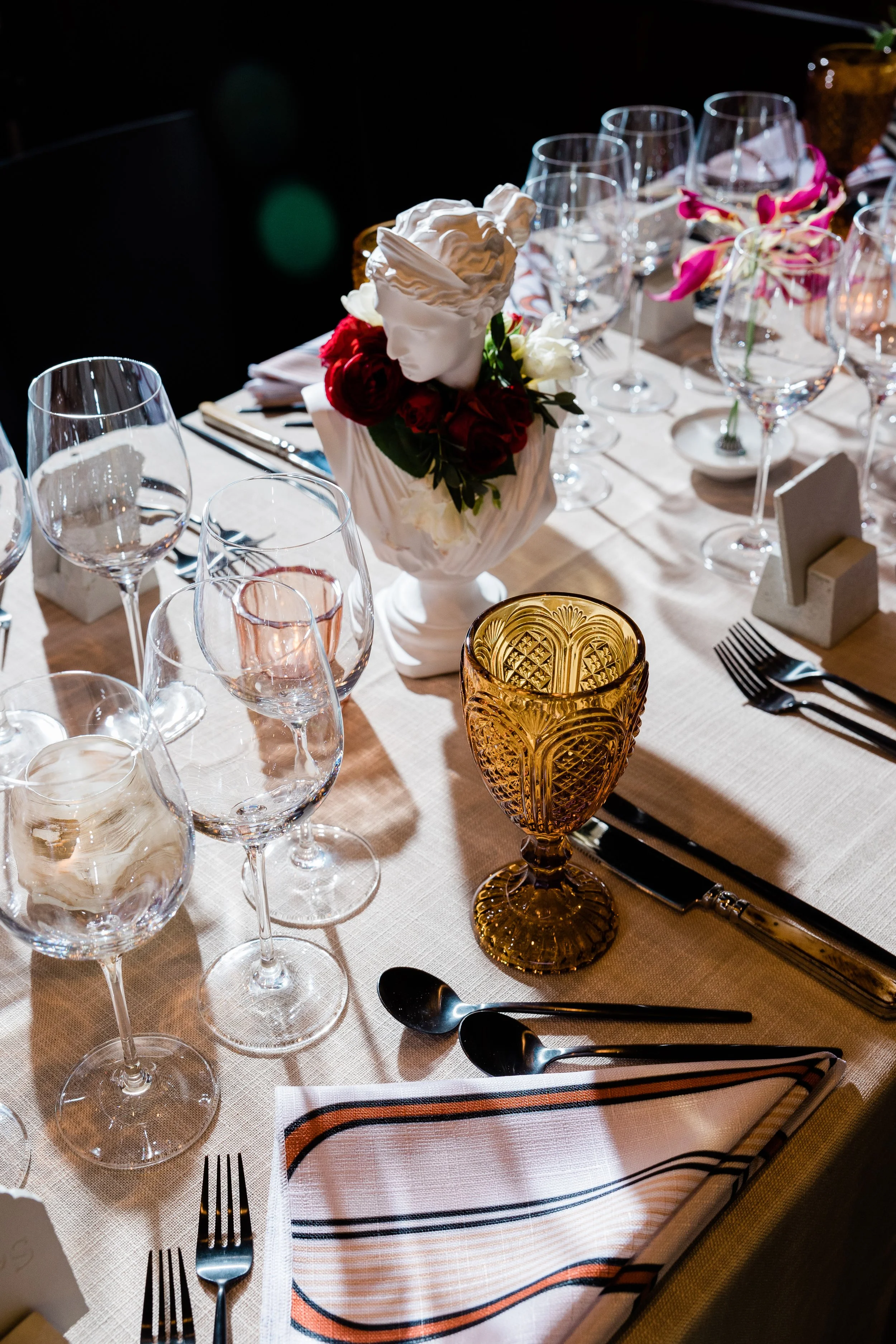 Elegant dining table set for a formal event with multiple wine glasses, a floral centerpiece, gold and black cutlery, and a wrapped napkin.