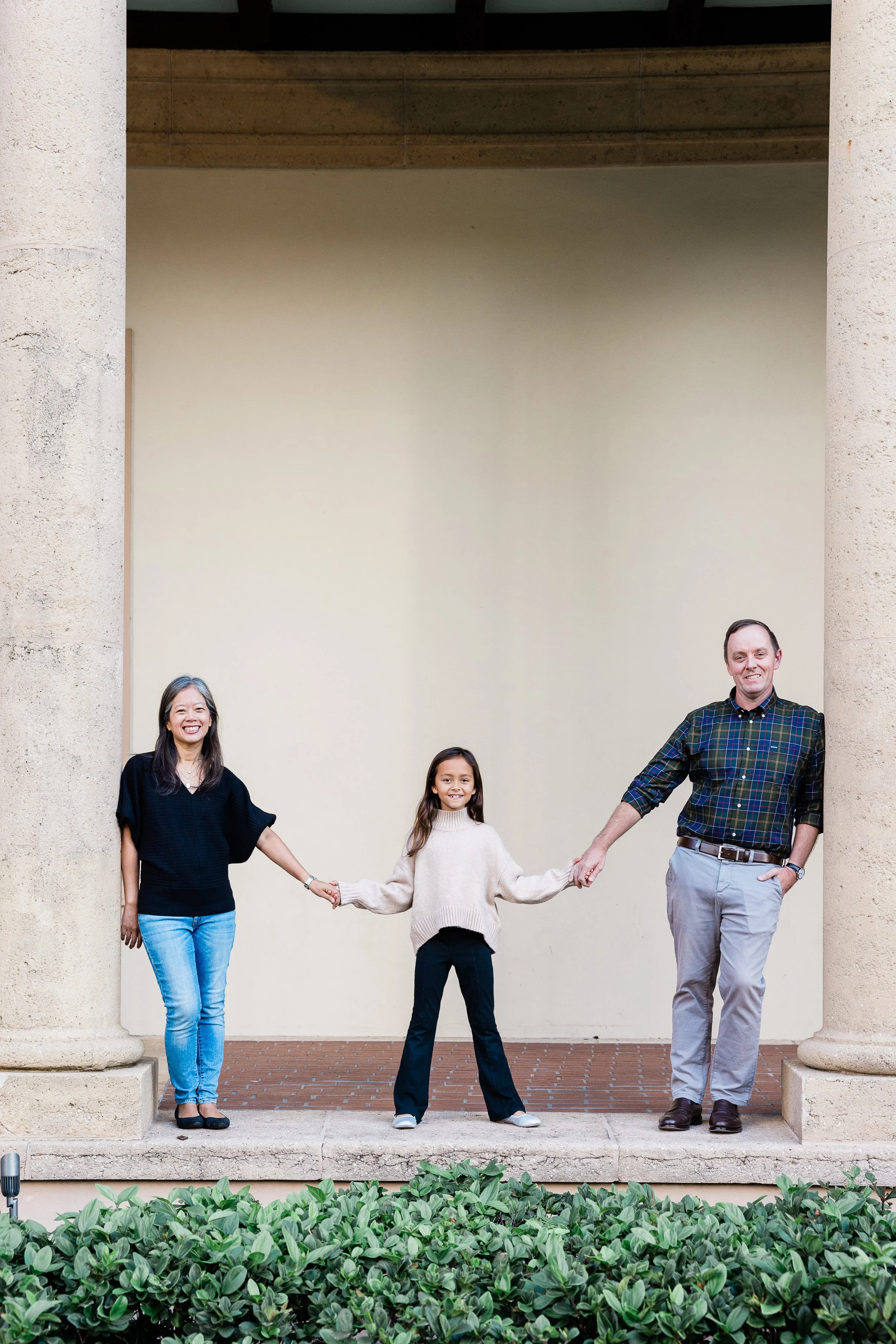 A family of four standing on a porch, holding hands in a line, smiling at the camera. The family includes a woman, a young girl, a teenage girl, and a man. The background features large columns and a beige wall, with green bushes at the bottom of the