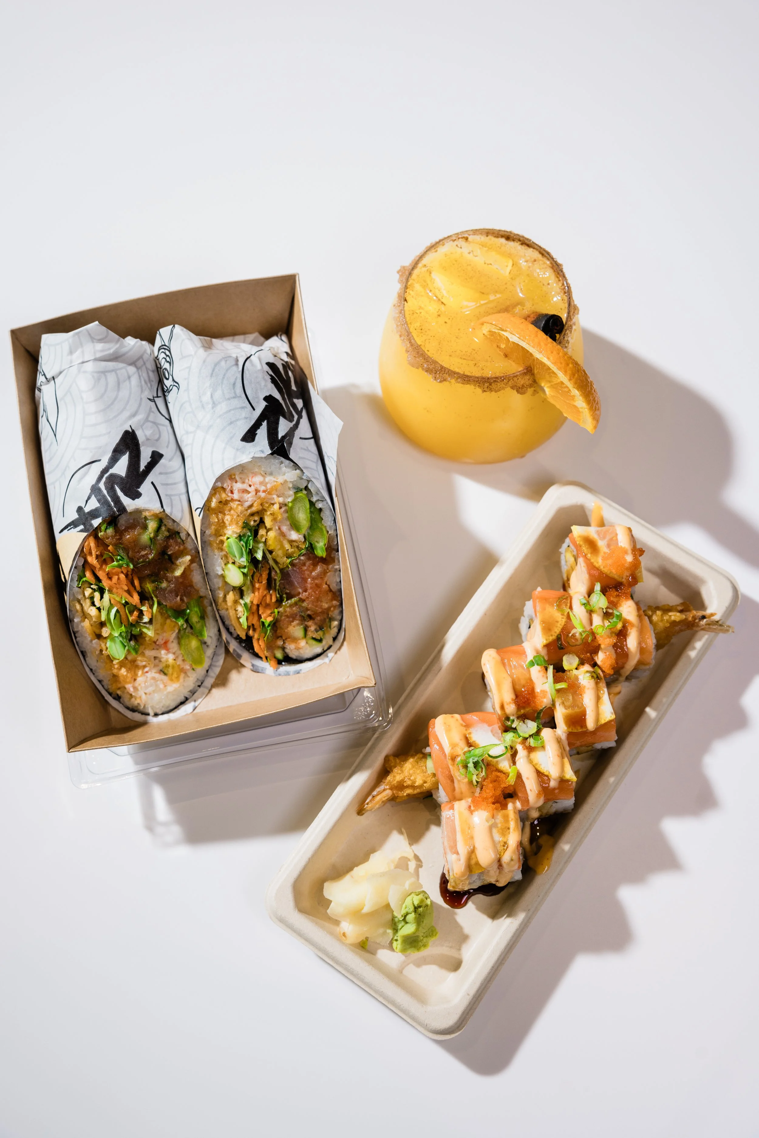 Three types of sushi with sauces, ginger, and wasabi, and a yellow cocktail with a sugar rim and orange garnish on a white surface.