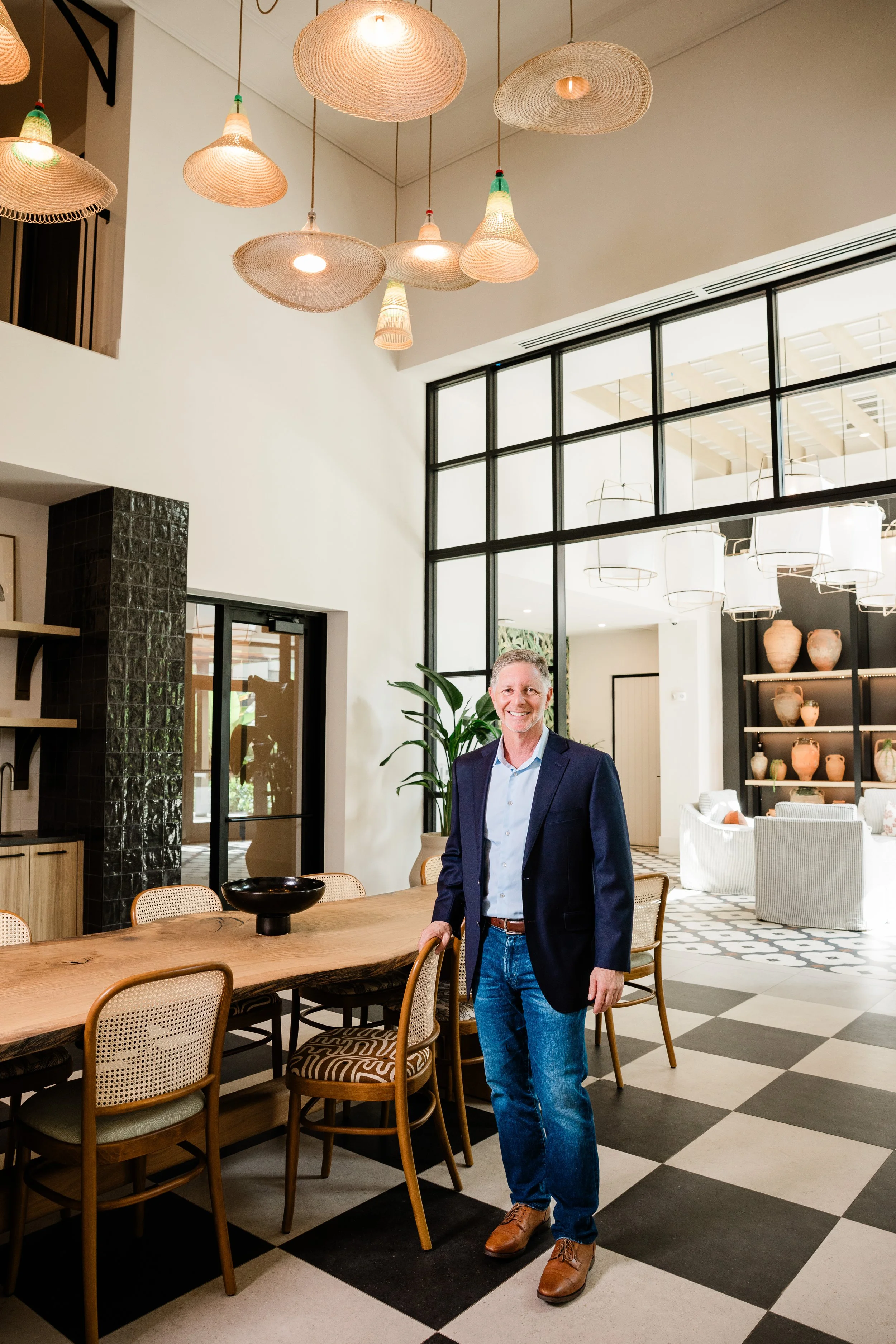 A man in a dark blazer, light blue shirt, and jeans standing in a modern dining room with black and white checkered flooring, a large wooden table, beige chairs, and a large window with black framing. The room has a high ceiling with various pendant 
