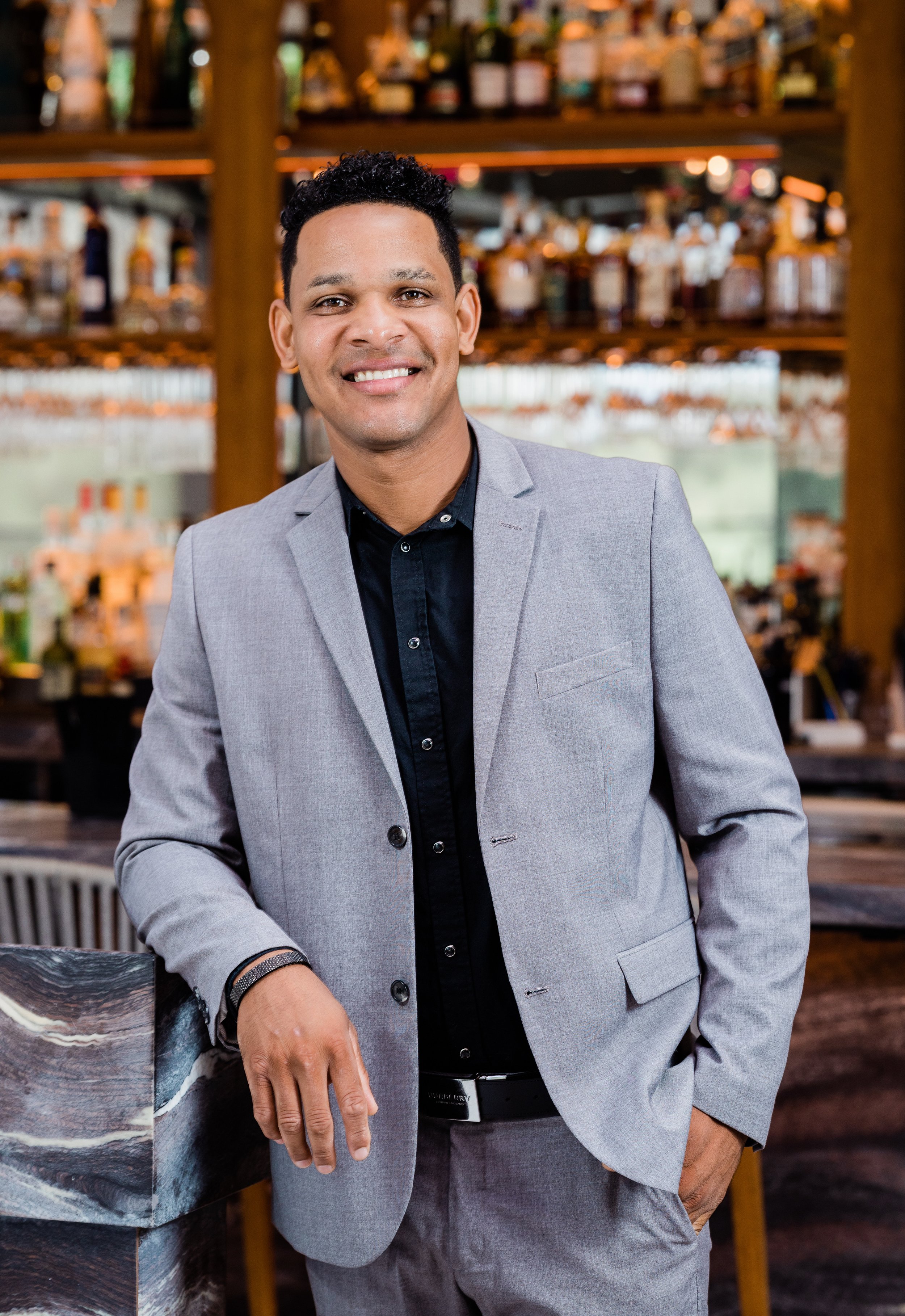 A man in a light gray suit standing in a bar or restaurant, smiling, with shelves of bottles behind him.