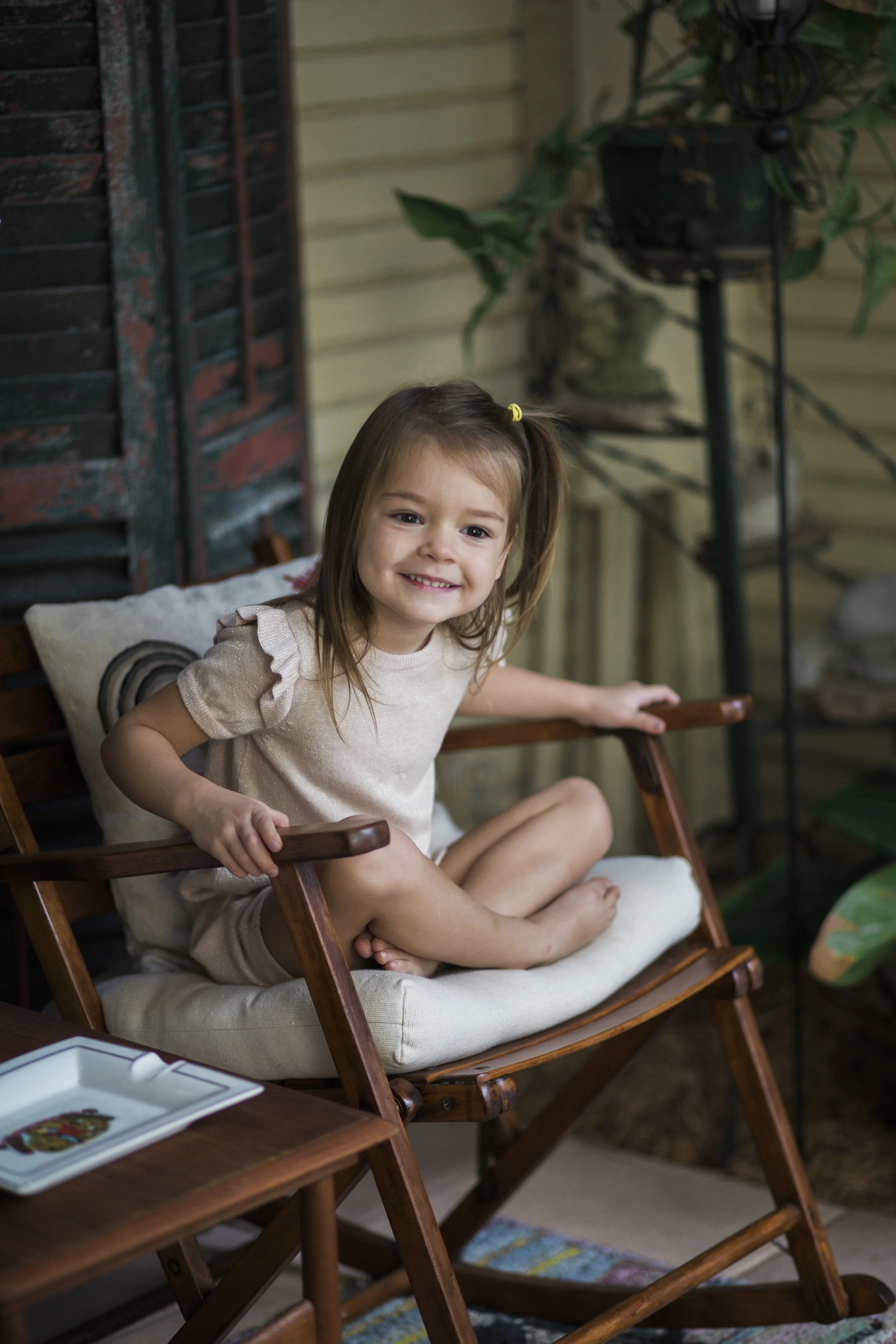 A young girl sitting in a wooden chair with a light-colored cushion, smiling at the camera. She is wearing a beige shirt with ruffle sleeves and has a small ponytail with a yellow hair tie. The setting appears to be a porch or backyard with plants an
