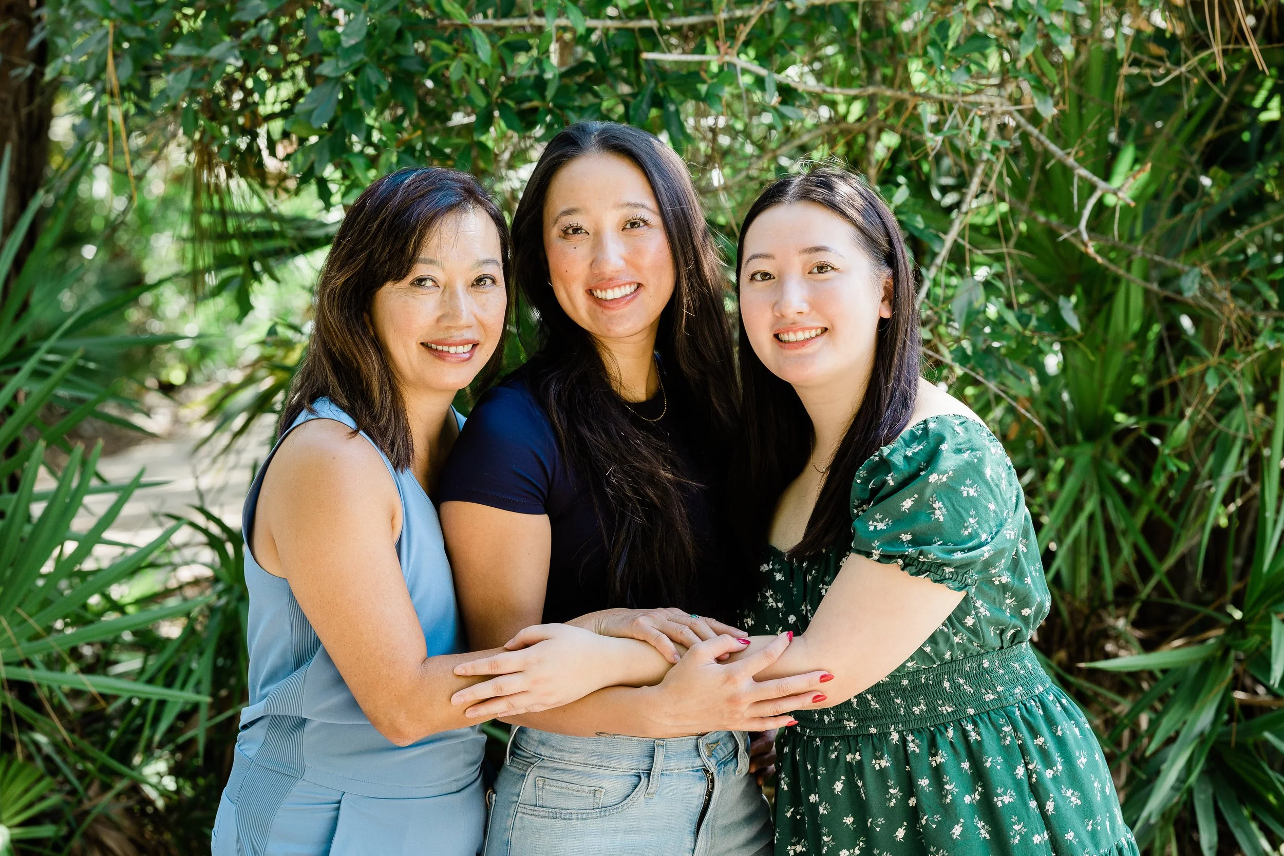 Three women with dark hair standing close together outdoors, smiling, surrounded by green foliage.