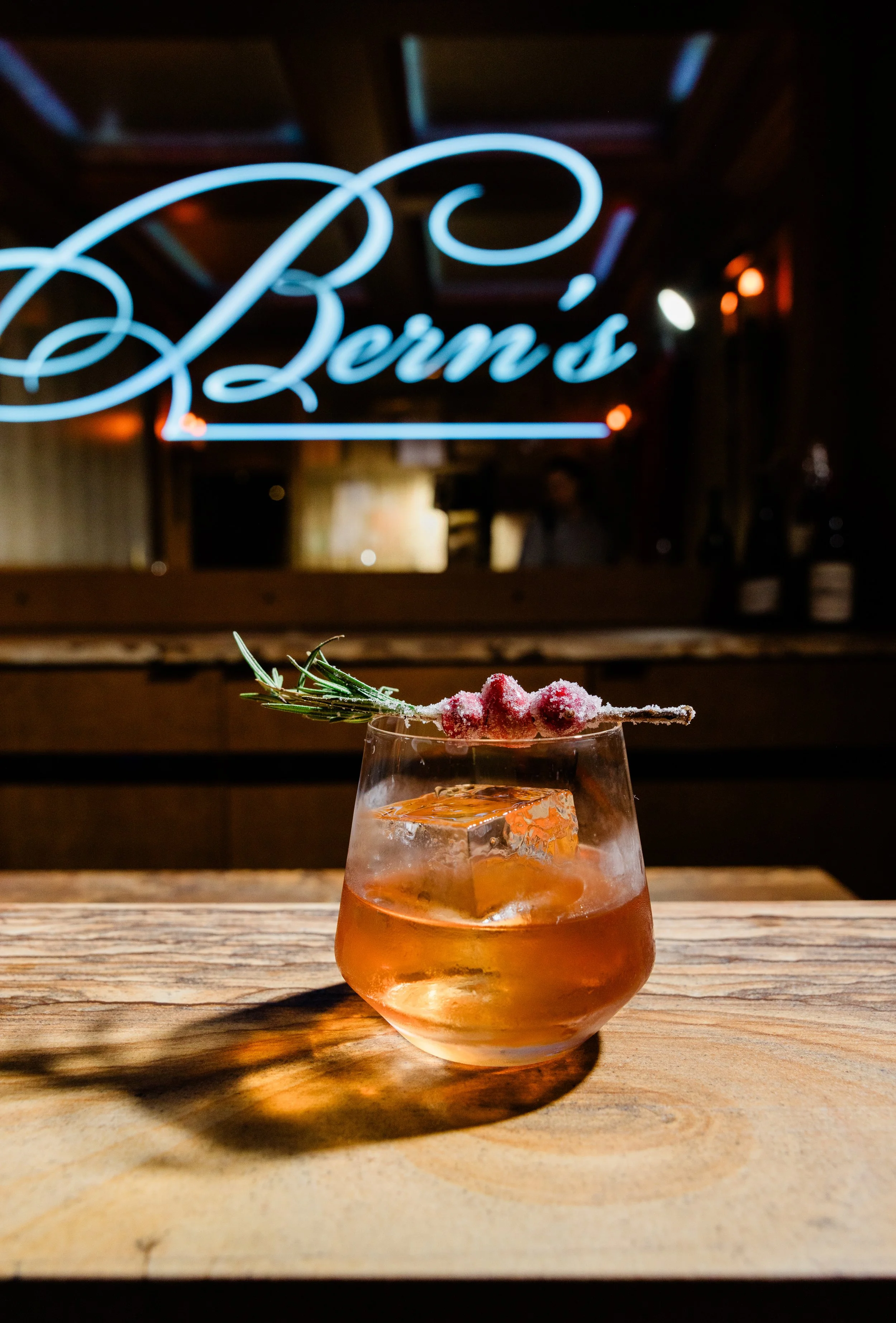 A glass of whiskey garnished with a sprig of rosemary and a sugared cranberries, sitting on a wooden table with a blurred neon sign that reads 'Bern's' in the background.