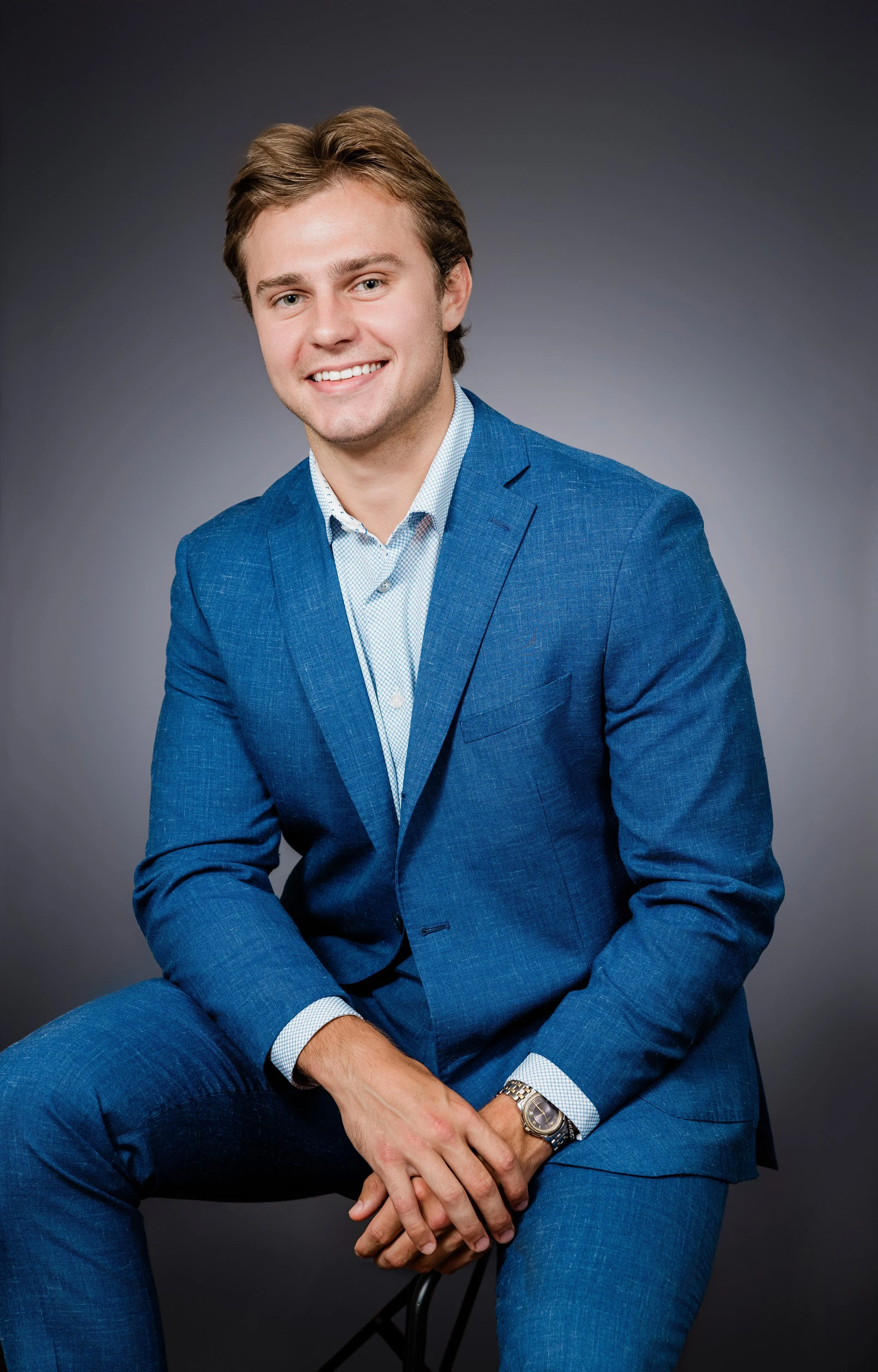 A young man with light brown hair, wearing a blue suit and a light blue dress shirt, is sitting against a dark gray background. He is smiling, with his hands resting on his lap, and is wearing a wristwatch on his left wrist.