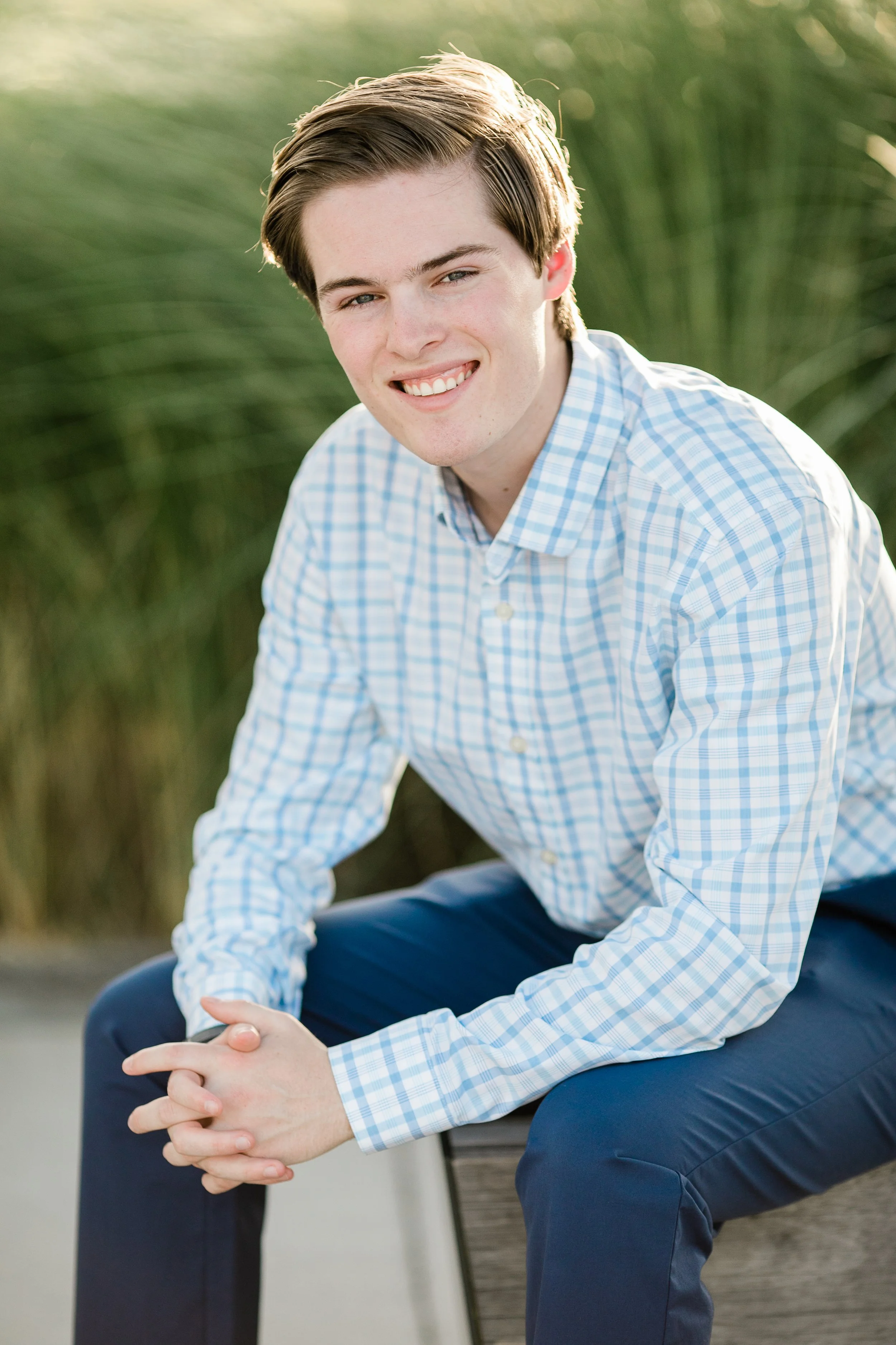 Young man sitting outdoors on a bench, smiling softly, wearing a light blue checkered shirt and dark pants, with greenery in the background.