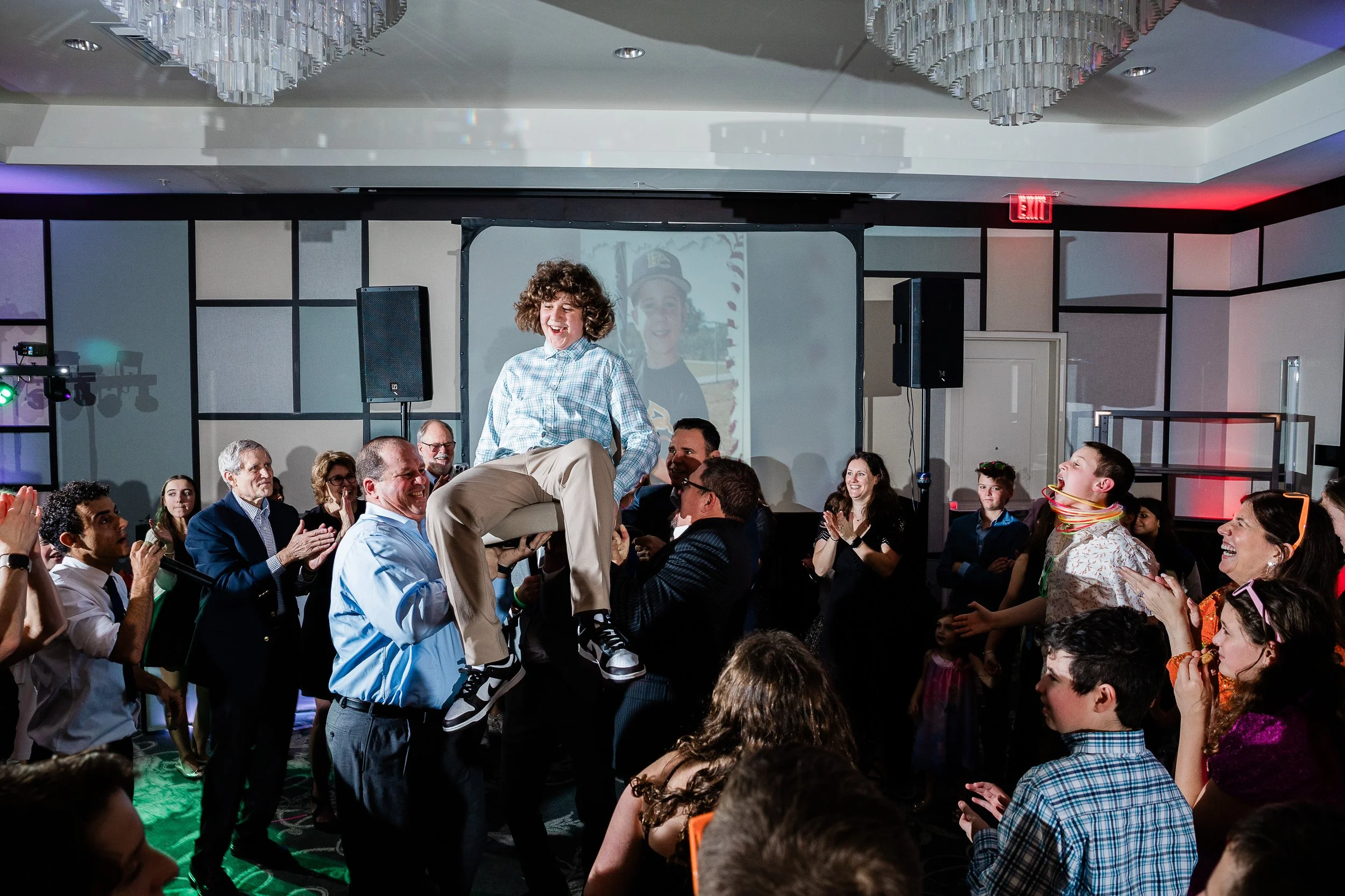 A lively indoor celebration scene with a group of people gathered around, where a young man is being lifted on someone's shoulders, smiling and laughing. The venue has a large chandelier and a screen in the background displaying a person’s image. Peo