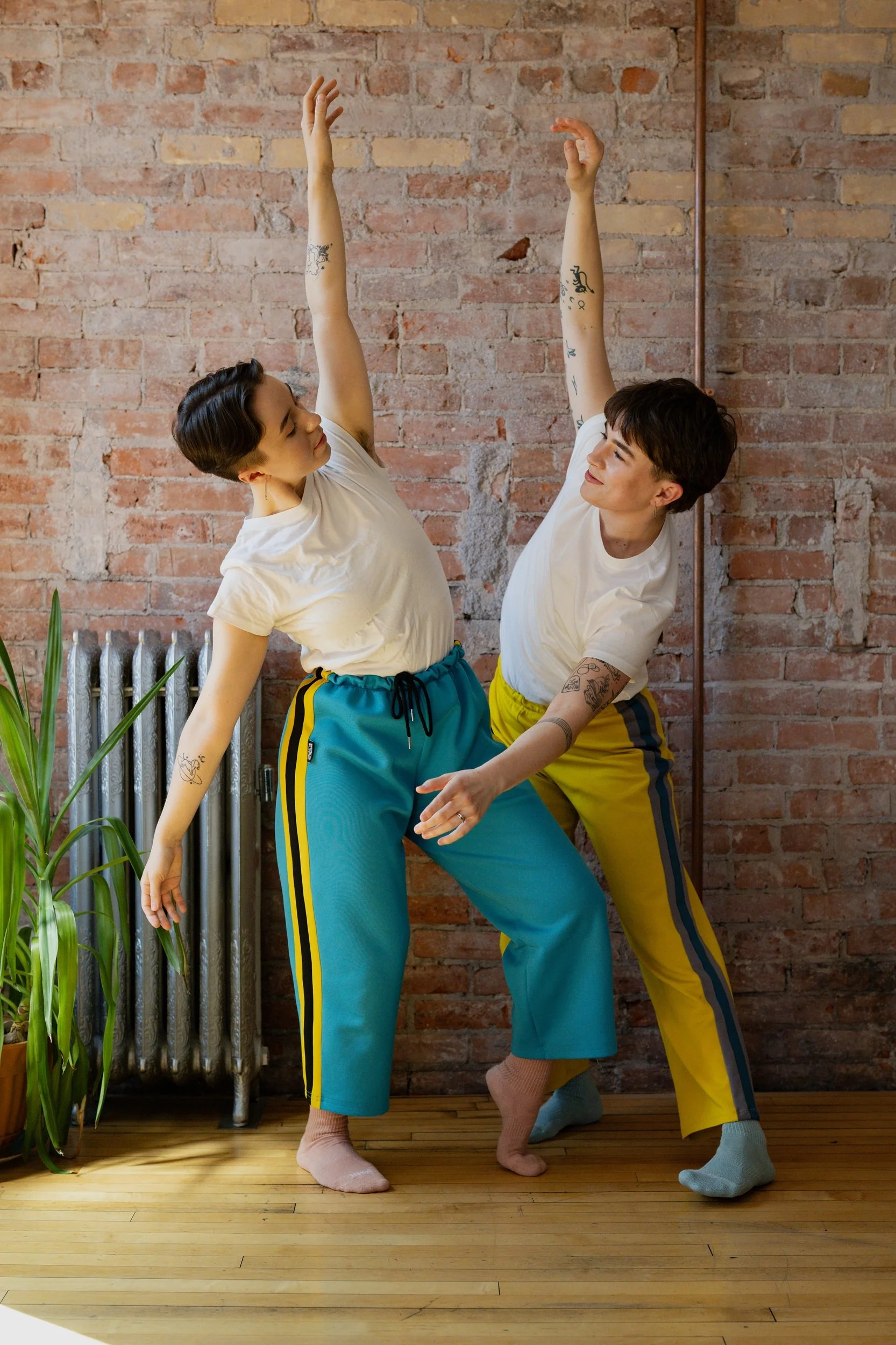 Two dancers with tattoos dance together indoors, raising their arms and smiling at each other, against a brick wall background.
