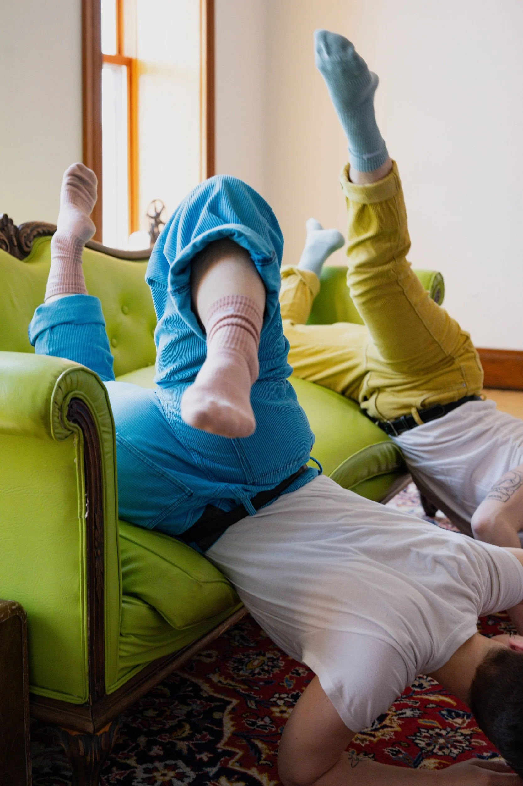 Two children are playing on a green vintage-style sofa. One child is wearing a hoodie and obscures their face with their arm, while the other is wearing yellow pants and socks, with their legs up in the air. The children are on a patterned red and black rug, with a window and beige walls in the background.