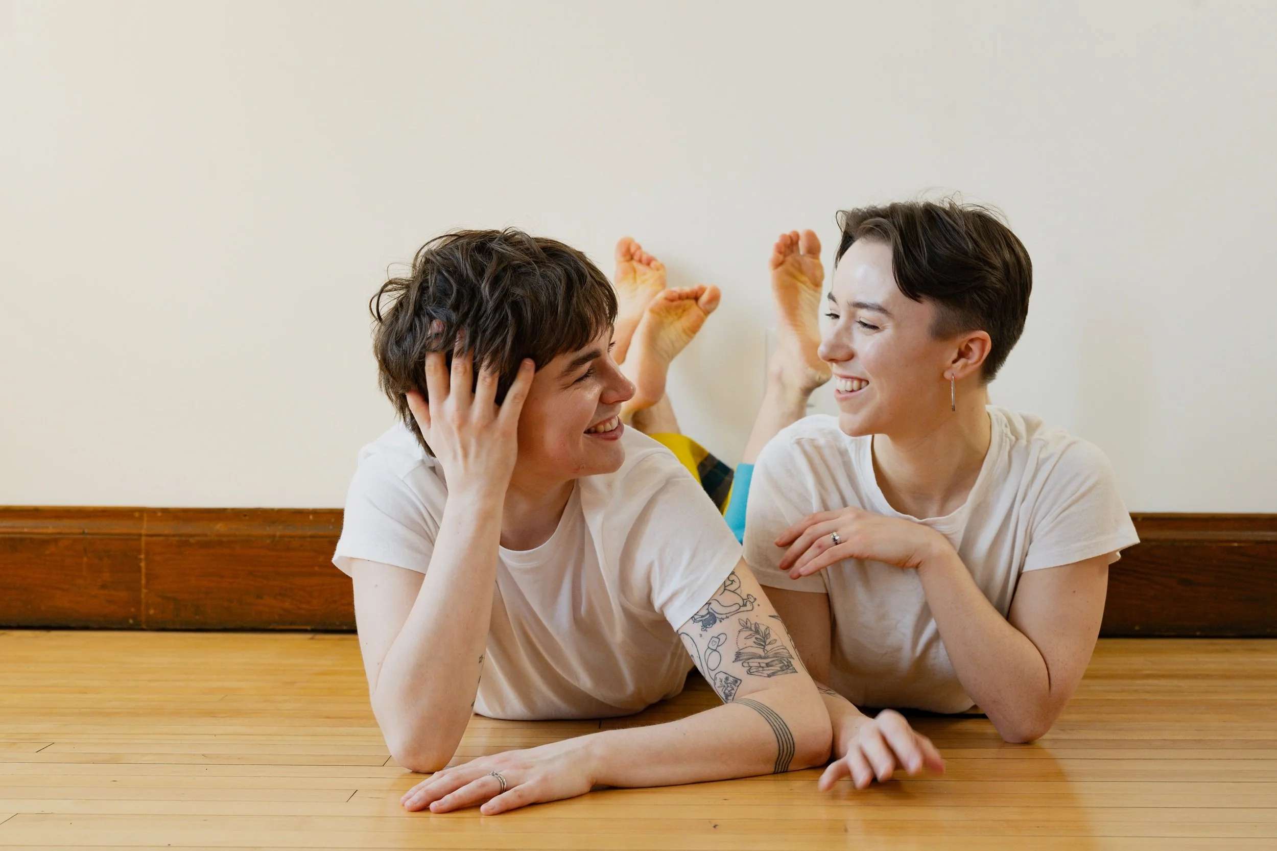 Two dancers are lying on their stomachs on a wooden floor, facing each other and smiling. One has short dark hair and the other has short wavy brown hair. They are indoors against a plain, light-colored wall.