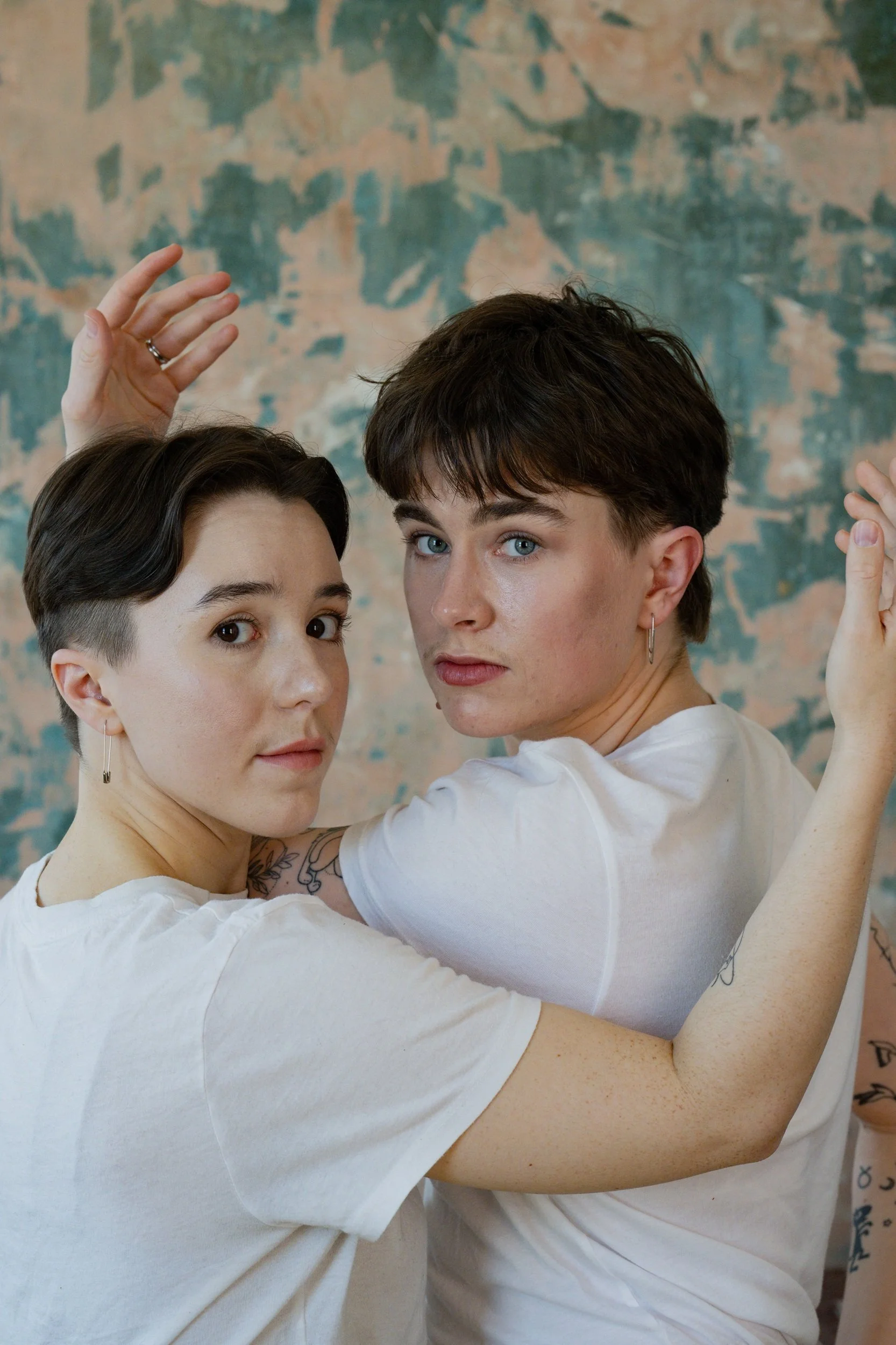 Two young adults, one with short dark hair and the other with short wavy brown hair, pose closely together with arms raised and intertwined, against a mottled blue and peach background.