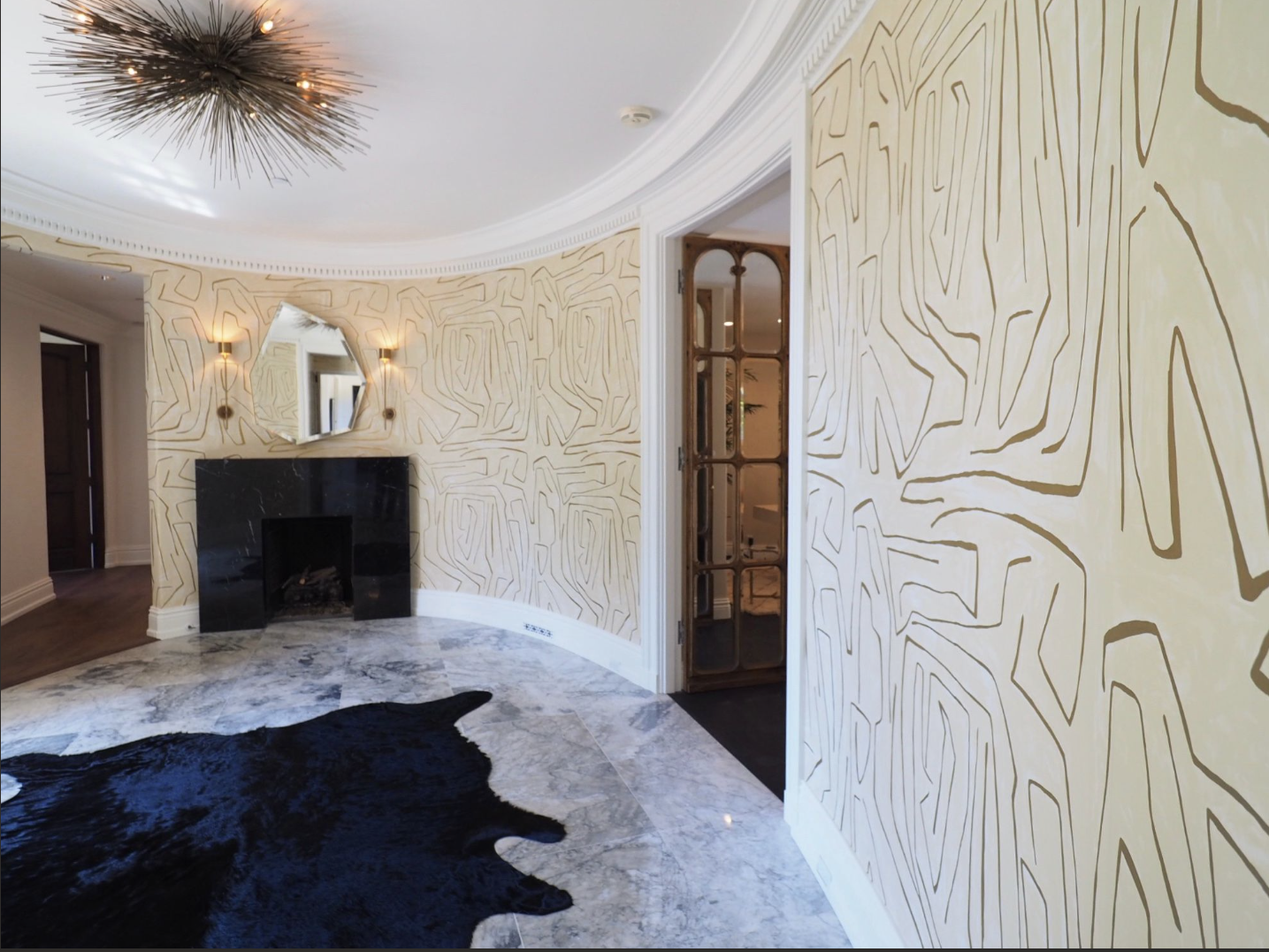 Interior of a room with modern design, featuring a black fireplace, marble flooring, patterned wallpaper, an irregular mirror, and a unique ceiling light fixture.