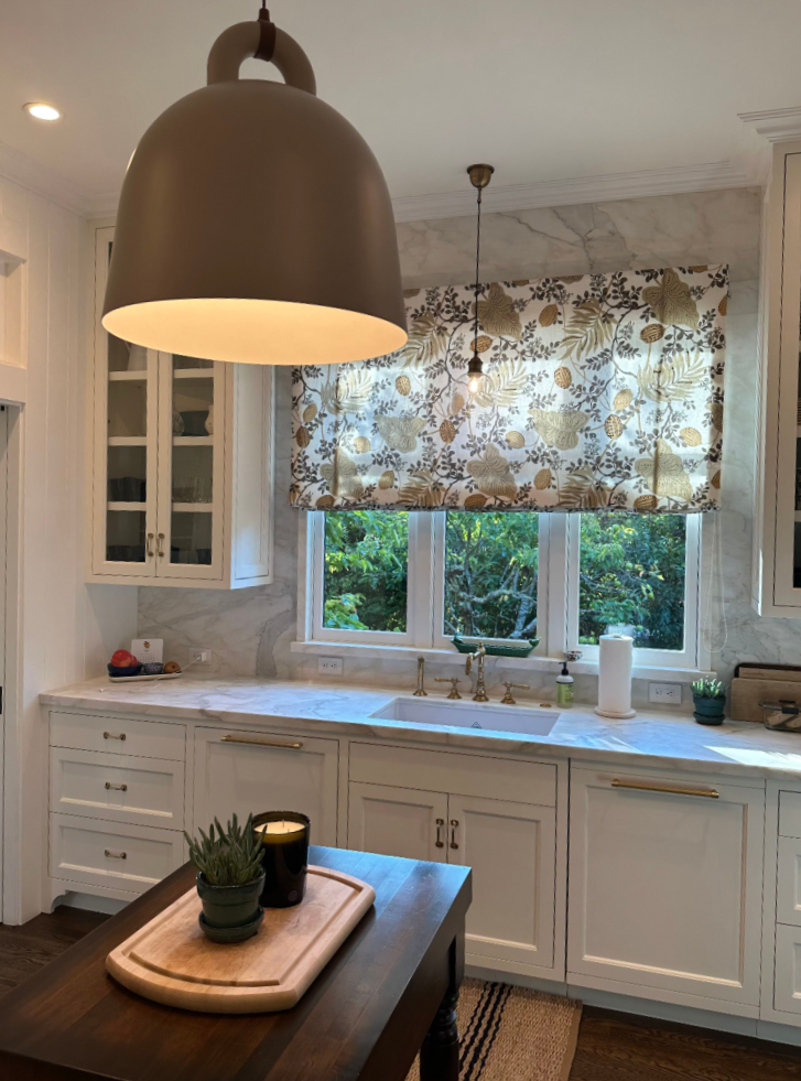 Kitchen with white cabinets, marble countertops, and a window with a floral valance. There is a large pendant light hanging above the kitchen island. On the countertop are various items including a soap dispenser, paper towel roll, and greenery. The window offers a view of green trees outside.