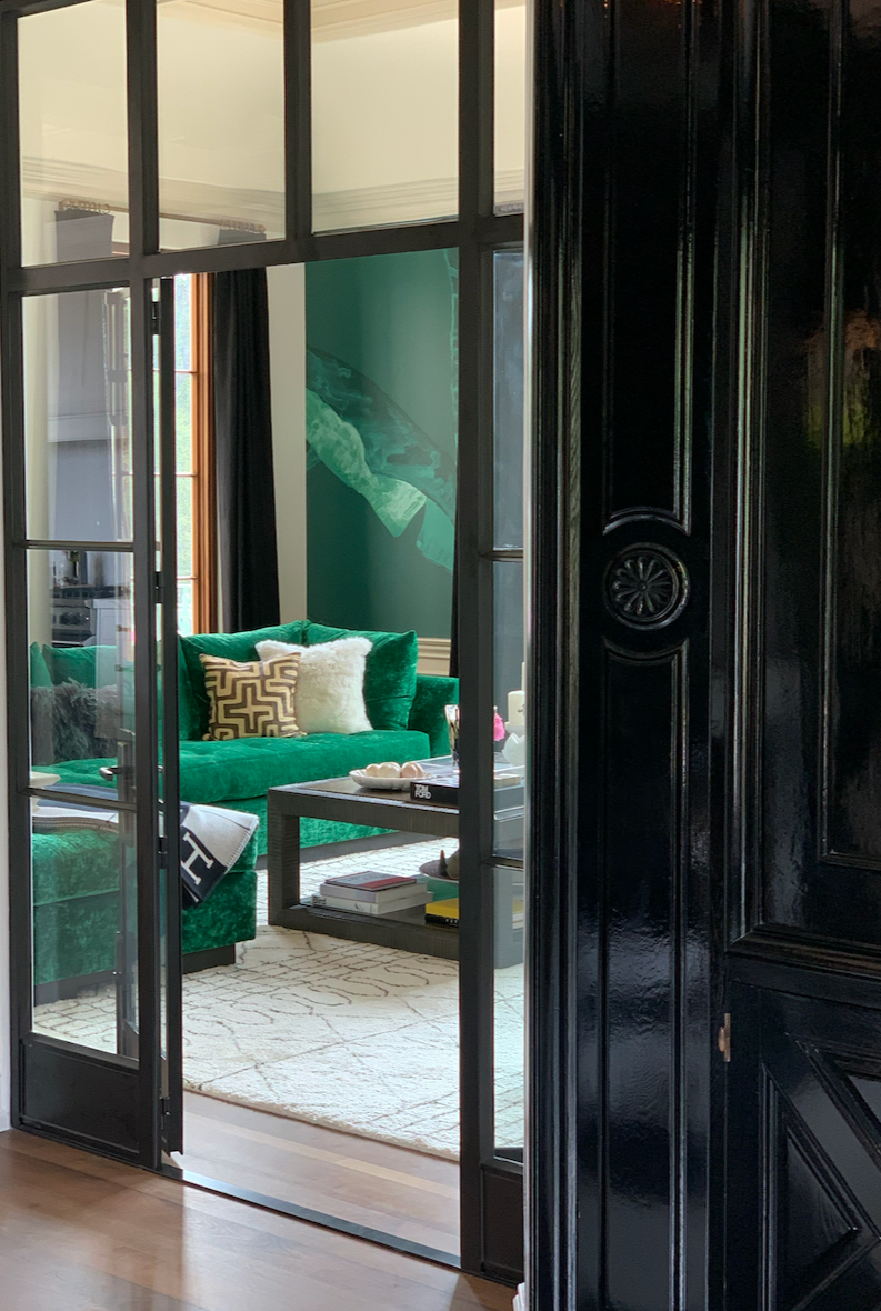View through glass doors into a living room with a green sofa, decorative pillows, a coffee table, and wall art blankets.