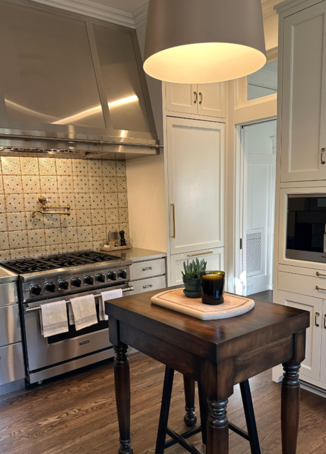 A modern kitchen featuring a stainless steel stove with a patterned backsplash, white cabinetry, a wooden table with a plant and cup, and a large pendant light overhead.