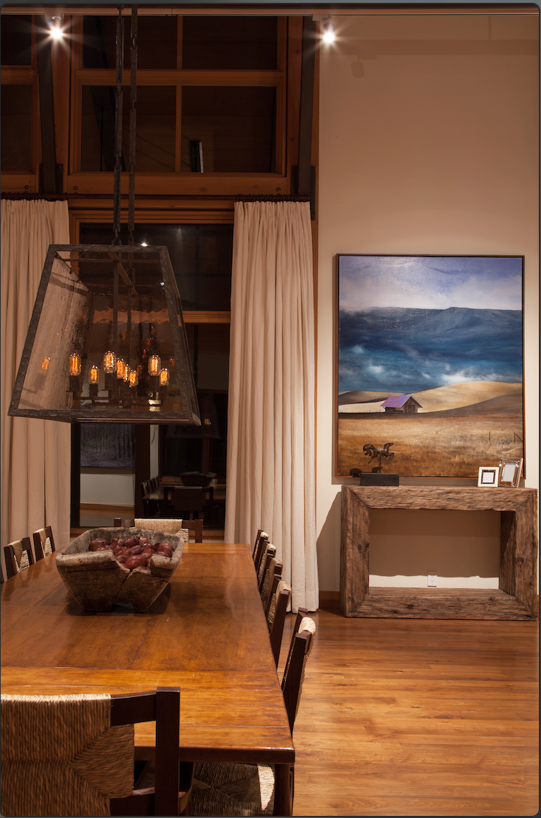 A dining room with a wooden table and chairs, a large bowl of onions, a chandelier with Edison bulbs, window with cream curtains, a landscape painting on the wall, and a wooden console table.
