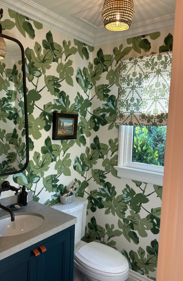 A small bathroom with leafy green patterned wallpaper, a window with a patterned shade, a toilet, and a blue vanity with a sink, black faucet, and a mirror.