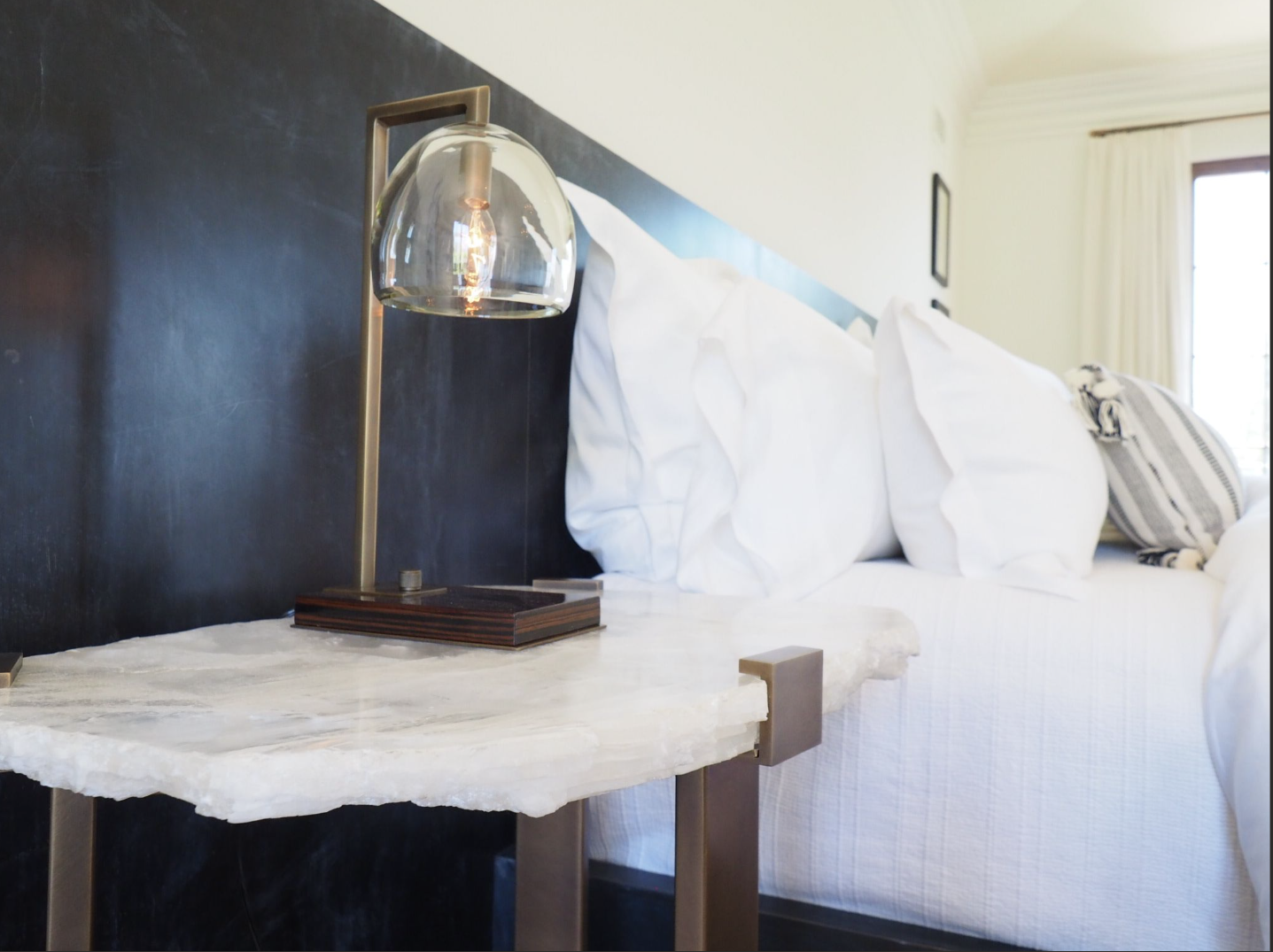 A bedside table with a modern glass table lamp next to a bed with white pillows and linens, in a well-lit room with a window and curtains.