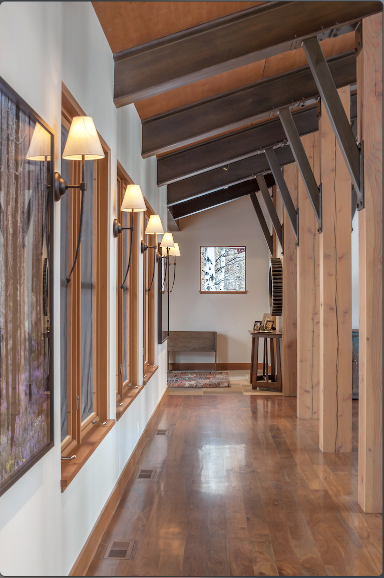 Interior view of a hallway with wood flooring, wood beams on the ceiling, wall-mounted lamps, and windows along one side.