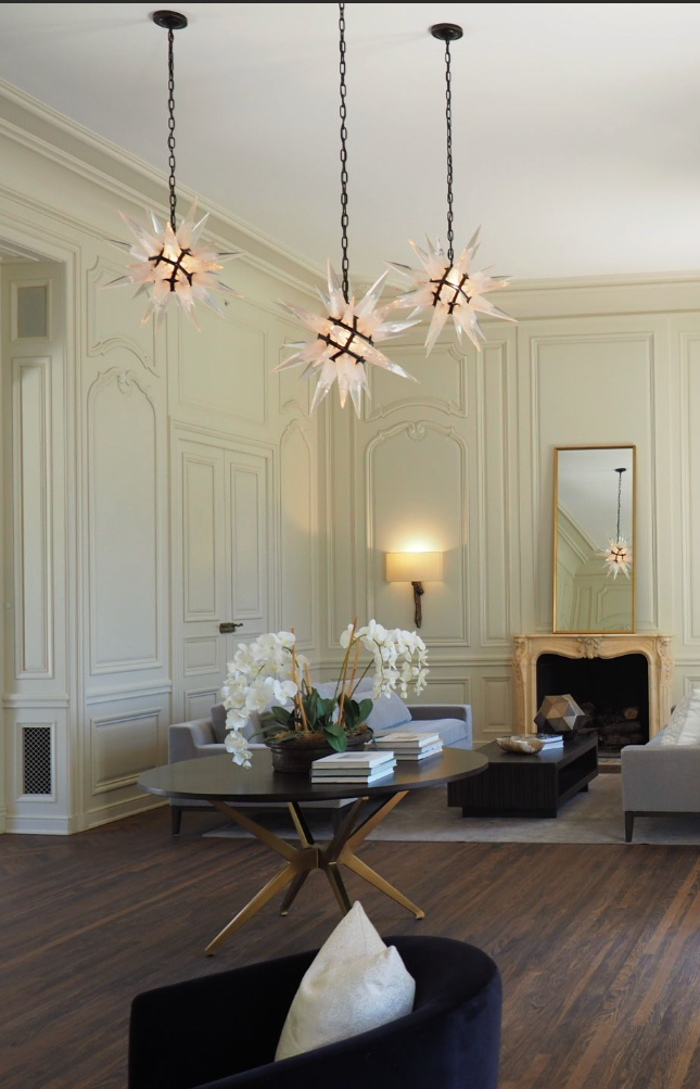 Modern living room with white paneled walls, a fireplace, a mirror above it, a wall sconce, and a table with white flowers and books. Hanging above are three starburst chandeliers with spiked glass design and black chains.