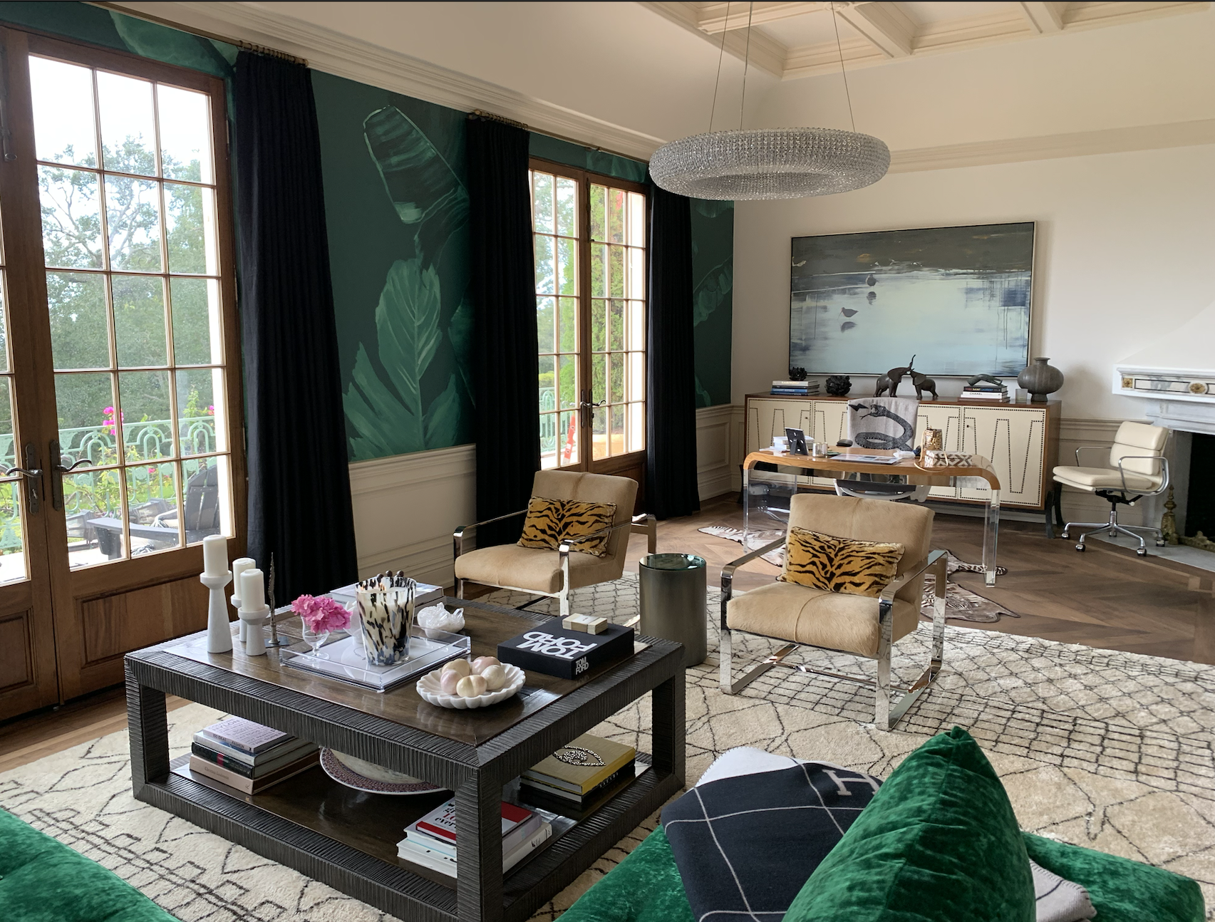 A stylish modern living room with large windows, beige armchairs with tiger-patterned pillows, a dark wooden coffee table with decorative items, a green velvet sofa, a patterned area rug, a white sideboard with artwork and decorative objects, and a glass chandelier hanging from the ceiling.