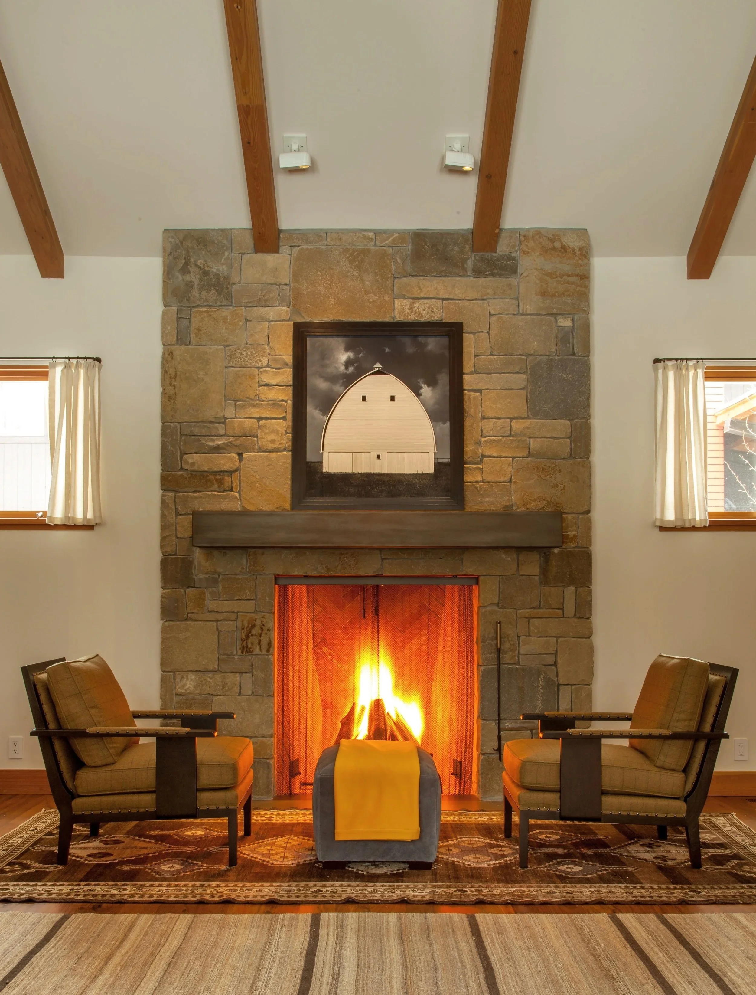 Living room with stone fireplace, two armchairs, a small ottoman, and a fire burning.