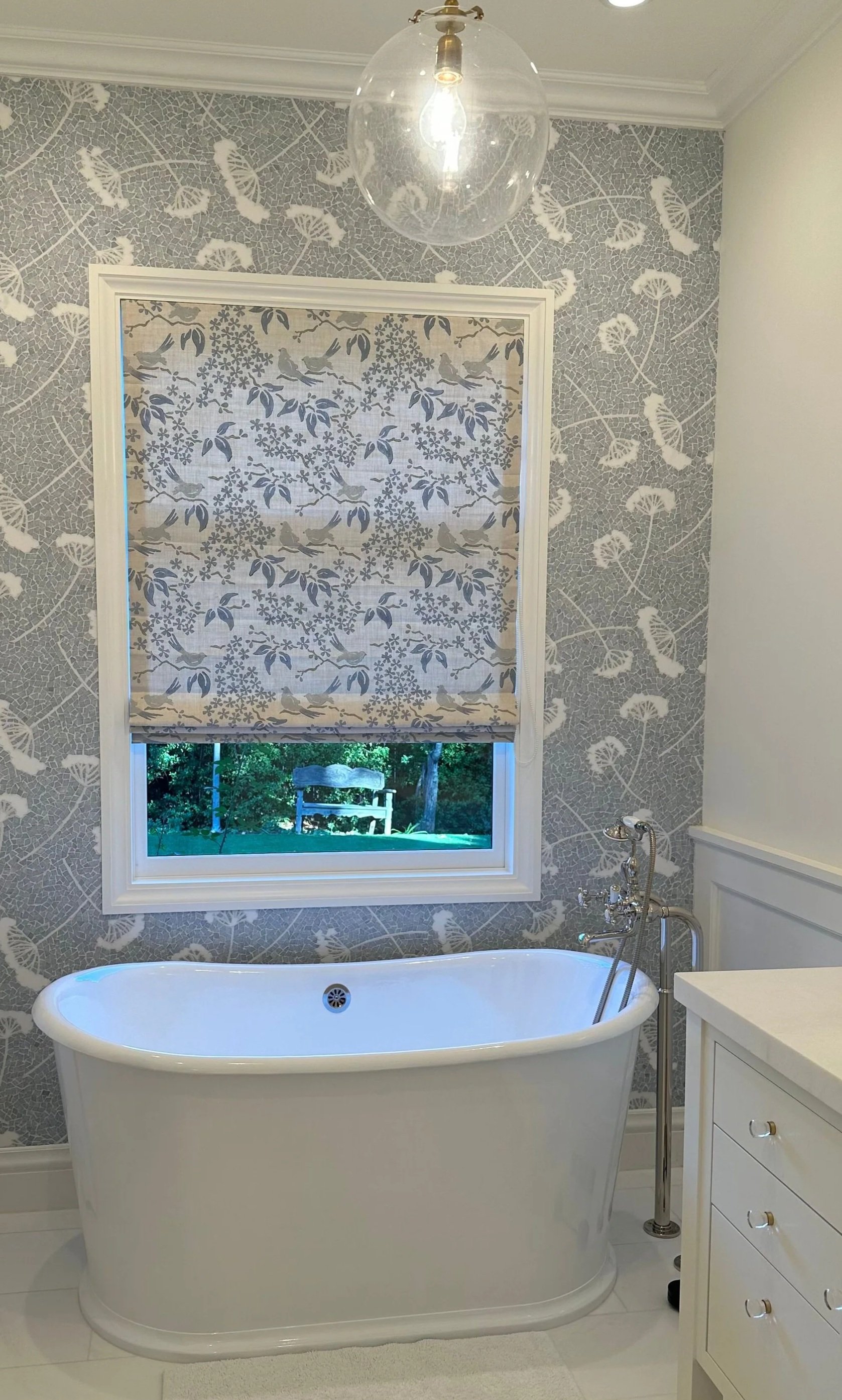 A bathroom with a white vintage bathtub, patterned wallpaper with bird and floral designs, a window with a decorative Roman shade, a glass pendant light, and a white vanity with drawers.