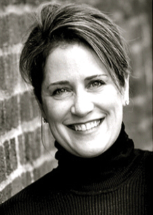 Black and white photo of a smiling woman with short hair, wearing a dark turtleneck, leaning against a brick wall.
