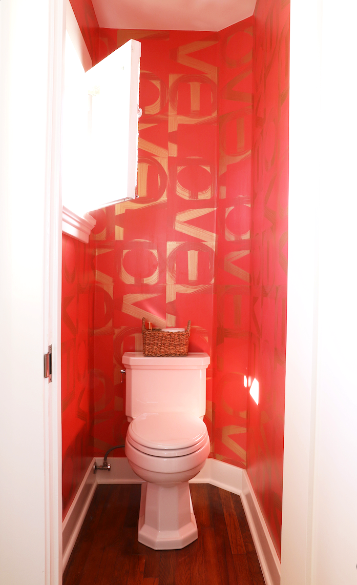 Small bathroom with red walls featuring large, bold, gold letters. There is a toilet with a woven basket on top and a small window on the left side, allowing natural light to brighten the space. The floor is wooden.