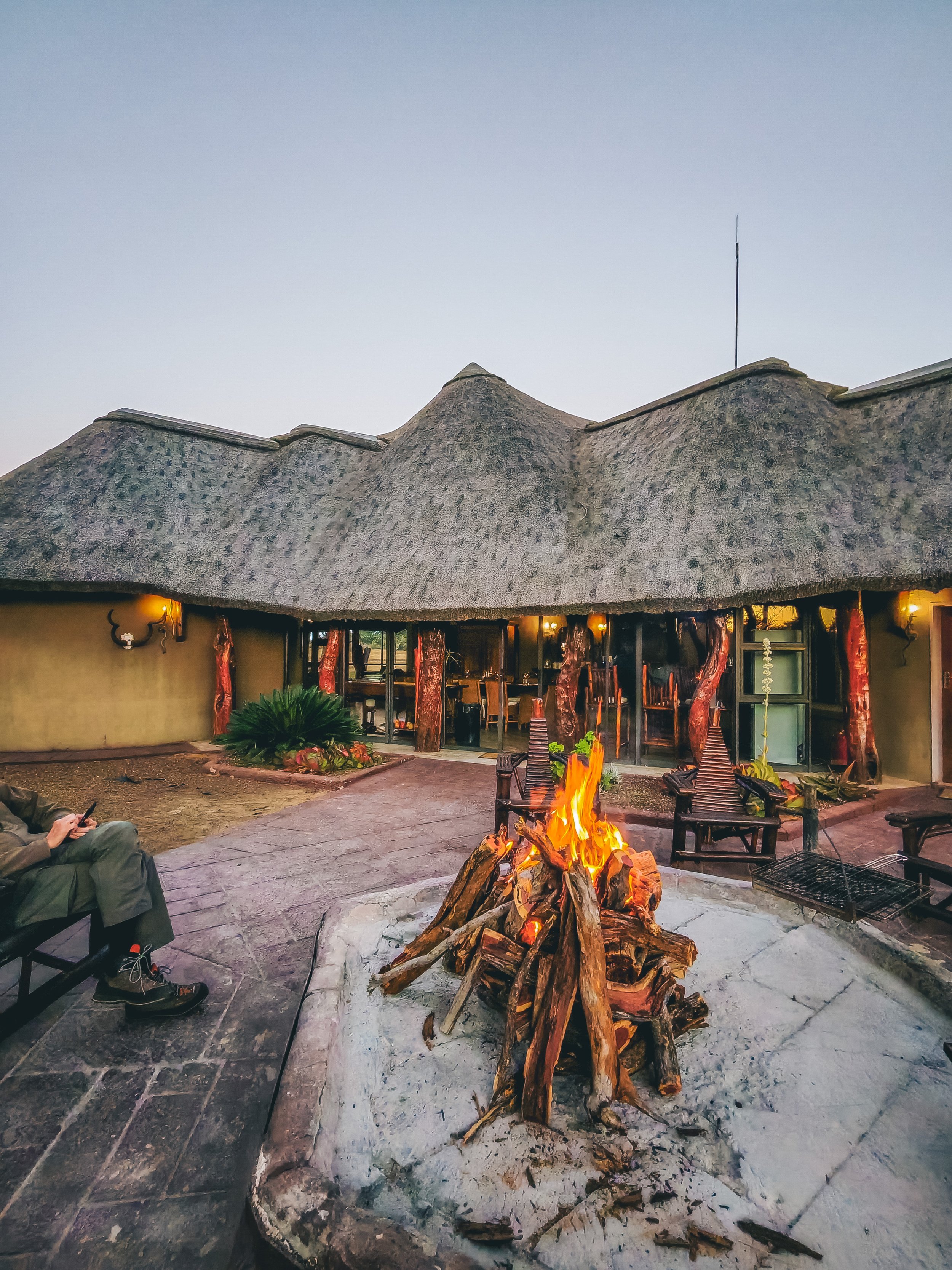 A person sitting outside near a stone fire pit with burning logs, facing a thatched-roof building at dusk, with outdoor seating visible.
