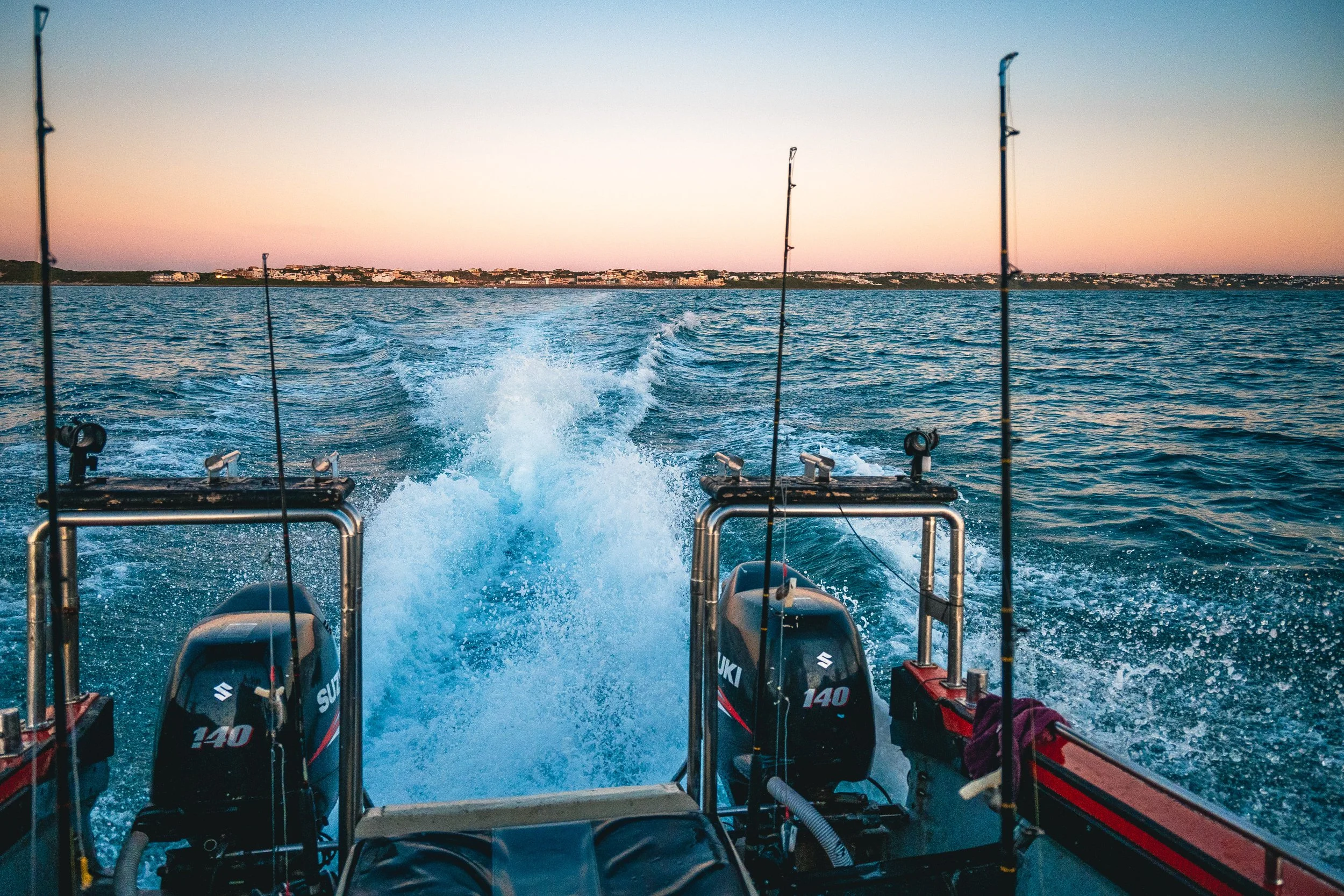 View from the back of a boat with two Suzuki 140 outboard motors, showing the wake in the water and fishing rods mounted on the boat, with a distant shoreline under a pastel-colored sunset sky.