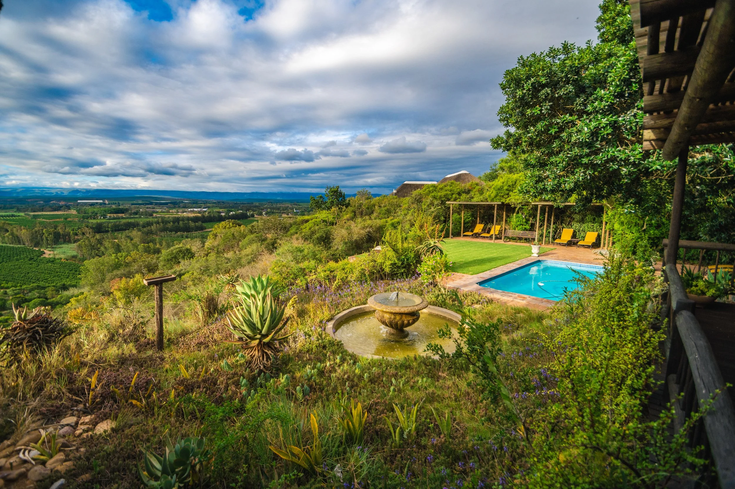 Scenic view of a backyard with a swimming pool, lounge chairs, a fountain, lush greenery, and a vast landscape with fields under a partly cloudy sky.