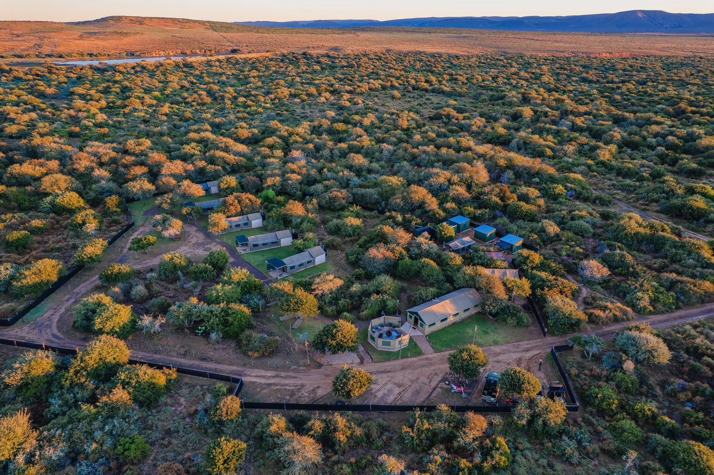 Aerial view of a rural property with multiple small houses, surrounded by trees and greenery, enclosed by a black fence, with dirt roads and vehicles visible, set in a semi-arid landscape with mountains in the background during sunset.