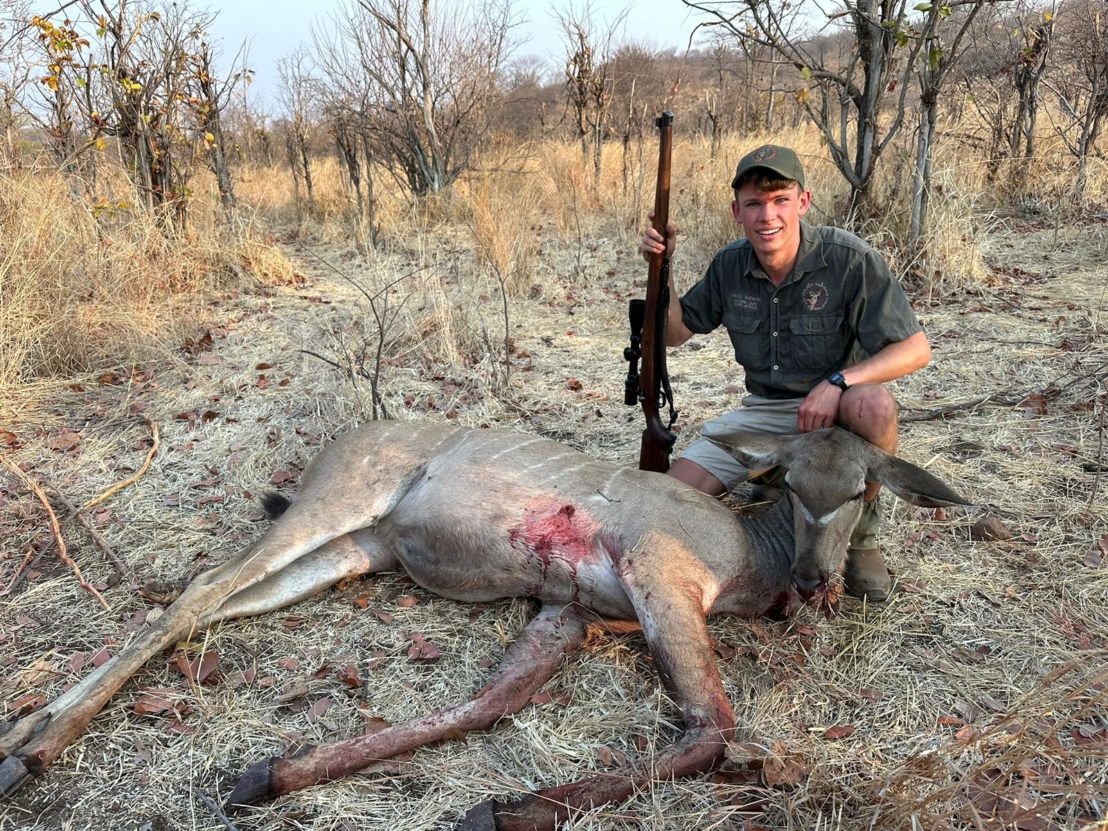 A young man dressed in outdoor clothing kneeling next to a dead deer with a rifle in the wilderness.