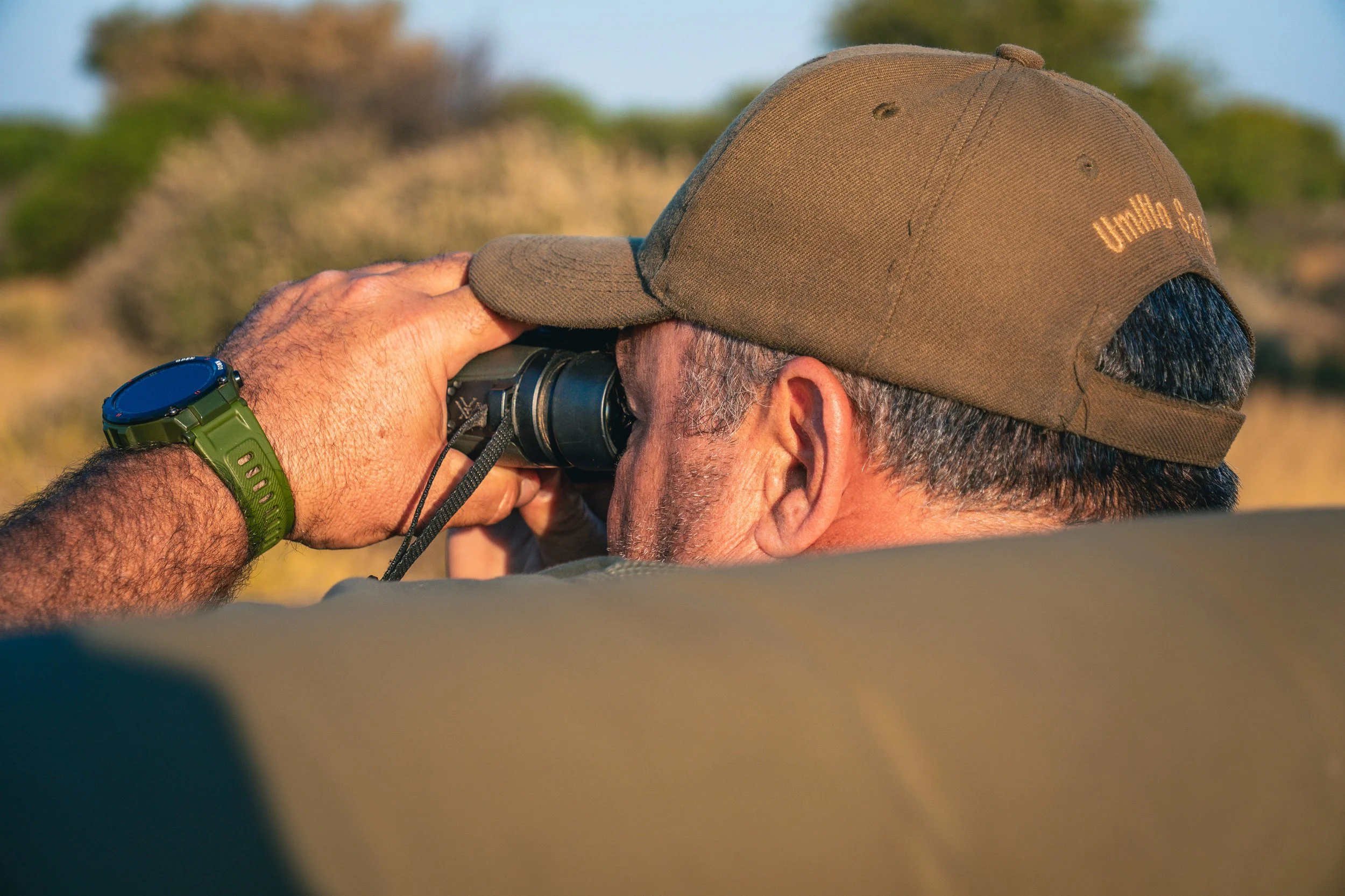 A man wearing a brown cap and a green watch is looking through binoculars outdoors during daylight.