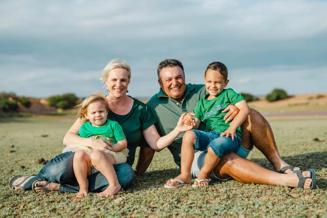 Smiling family of four sitting outdoors on the grass, with two children and two adults, holding hands and enjoying a sunny day.