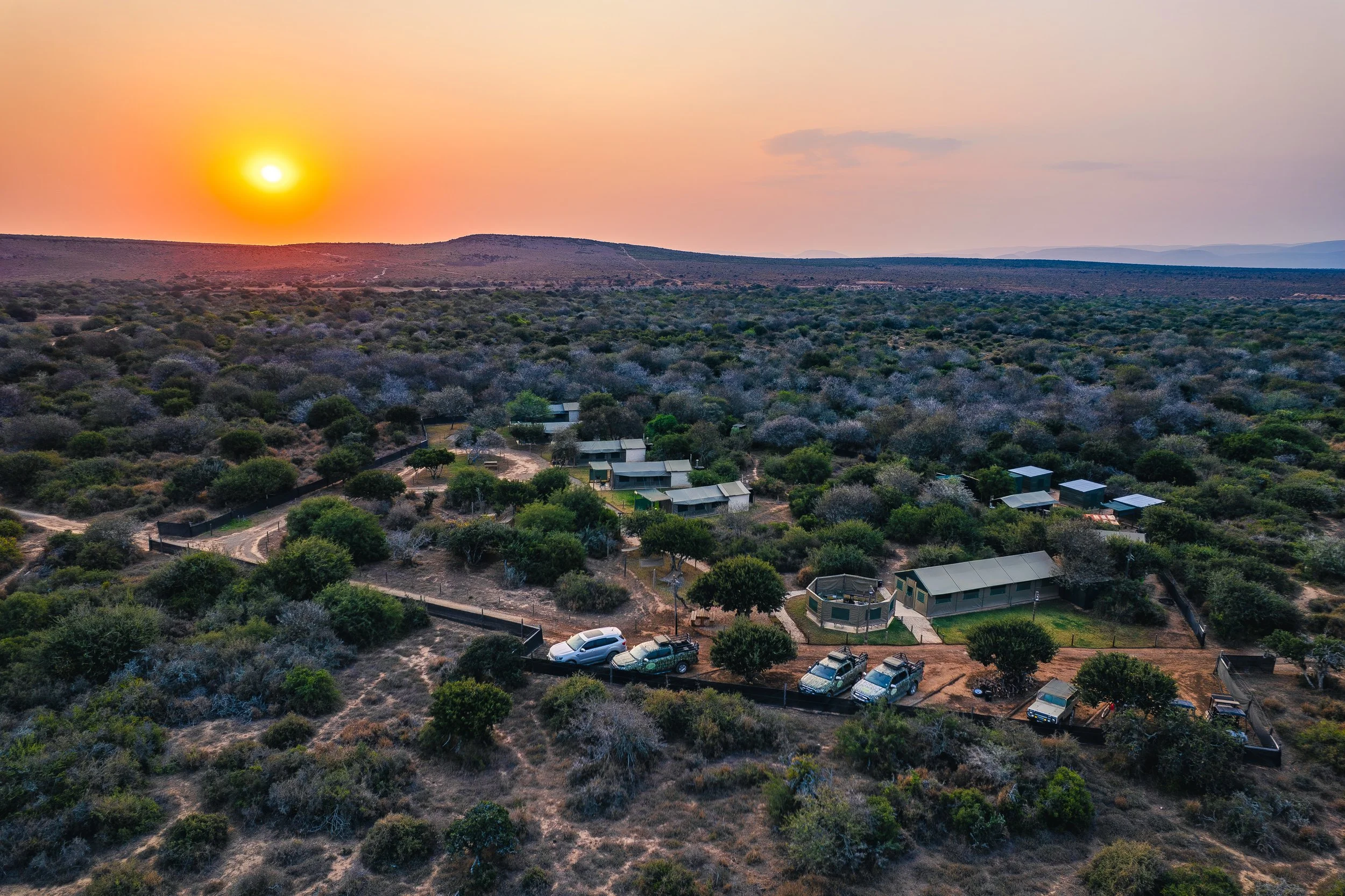 Sunset over a rural area with small buildings, trees, and a few parked cars.