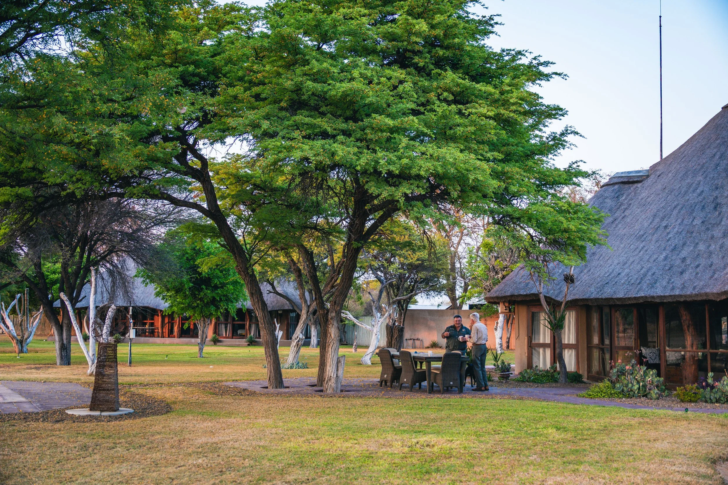 Two men standing and talking outside under trees near a thatched-roof building with outdoor furniture, green grass, and landscaping.