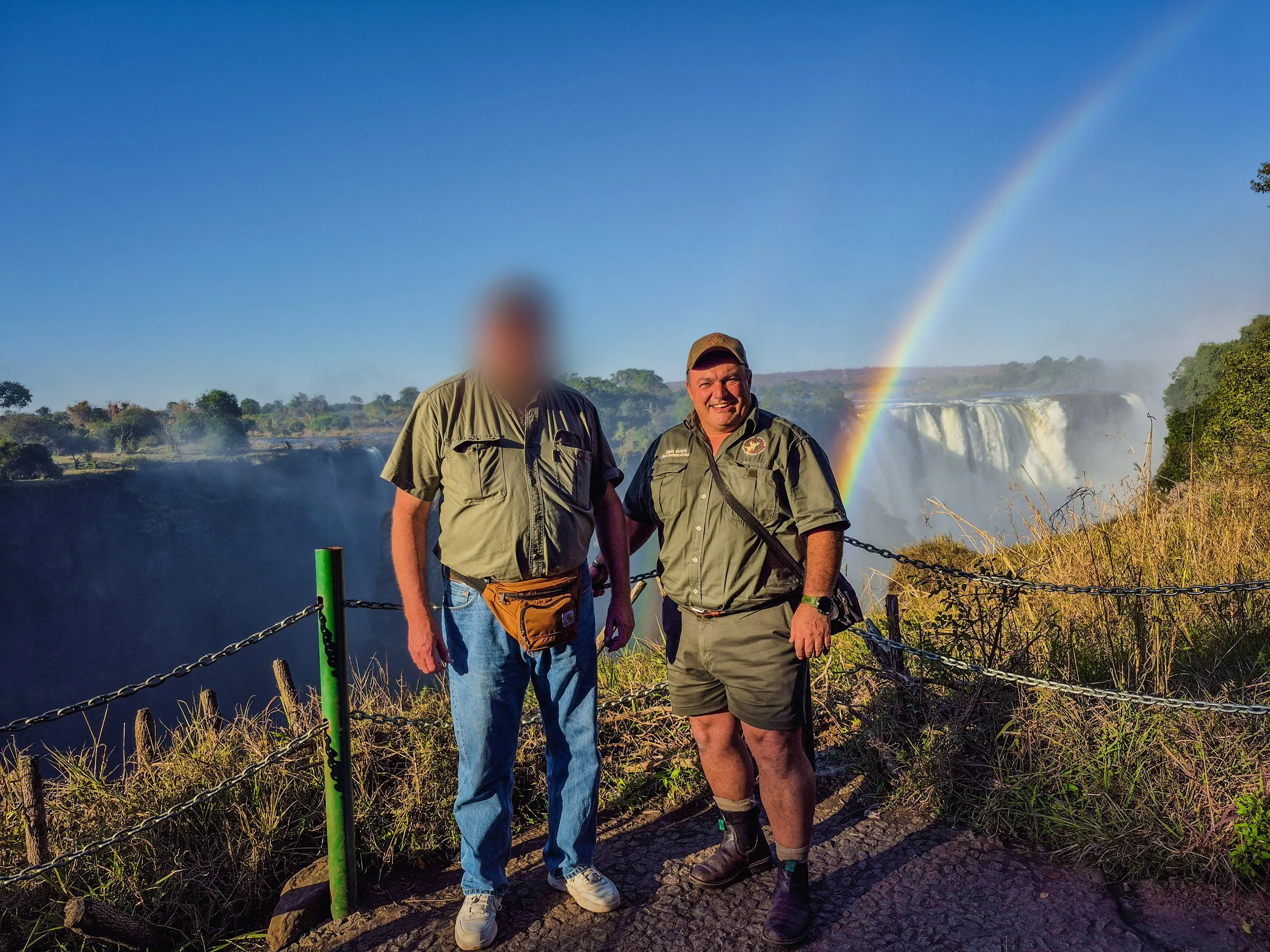 Two men standing on a viewing platform at a waterfall with a rainbow in the sky behind them.