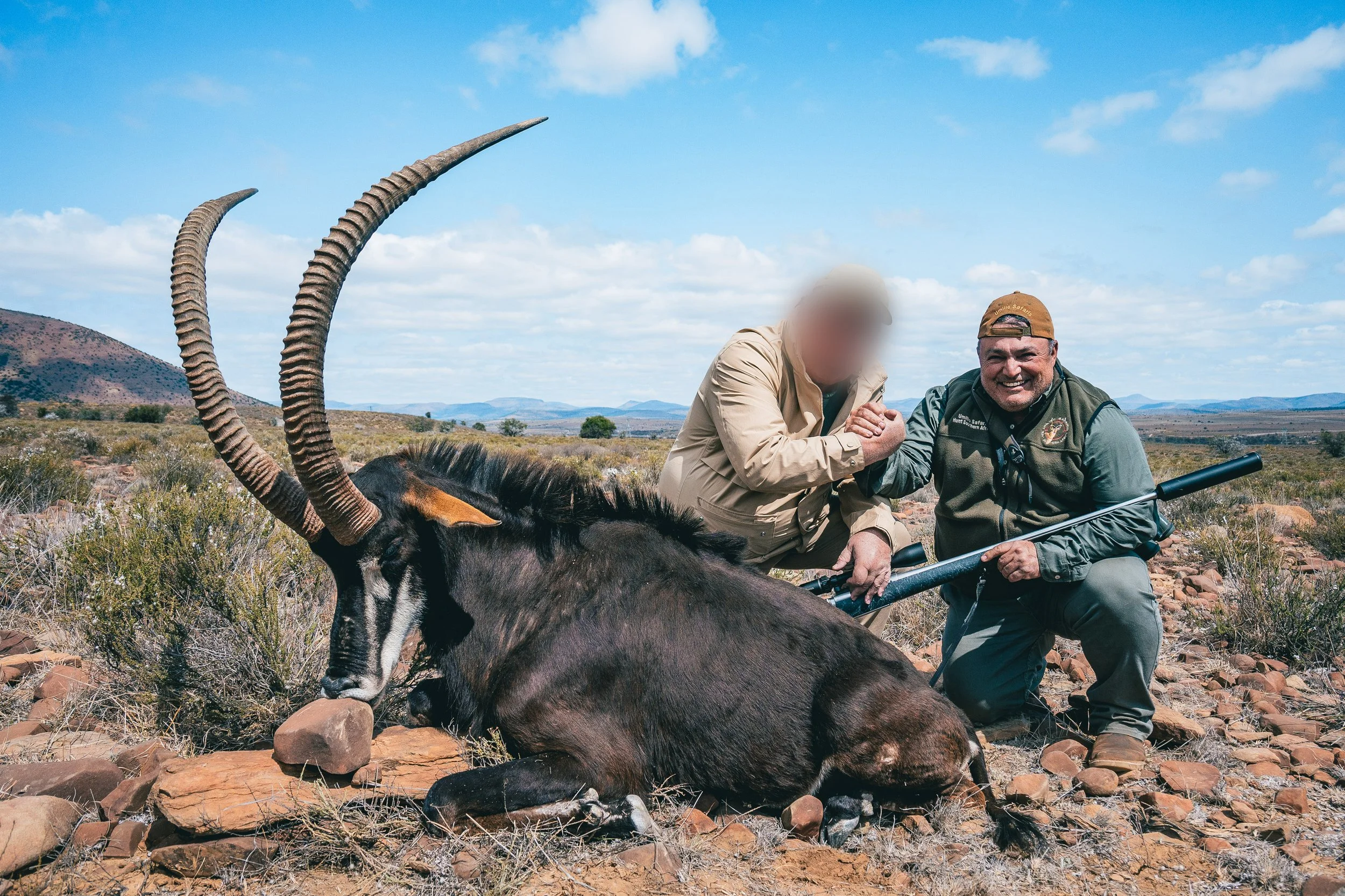 Two men kneel on rocky desert terrain next to a dead oryx antelope, which has long curved horns and is lying on the ground. One man is blurred, and the other is smiling; they are holding hands. The landscape features sparse vegetation, distant hills, and a blue sky with clouds.