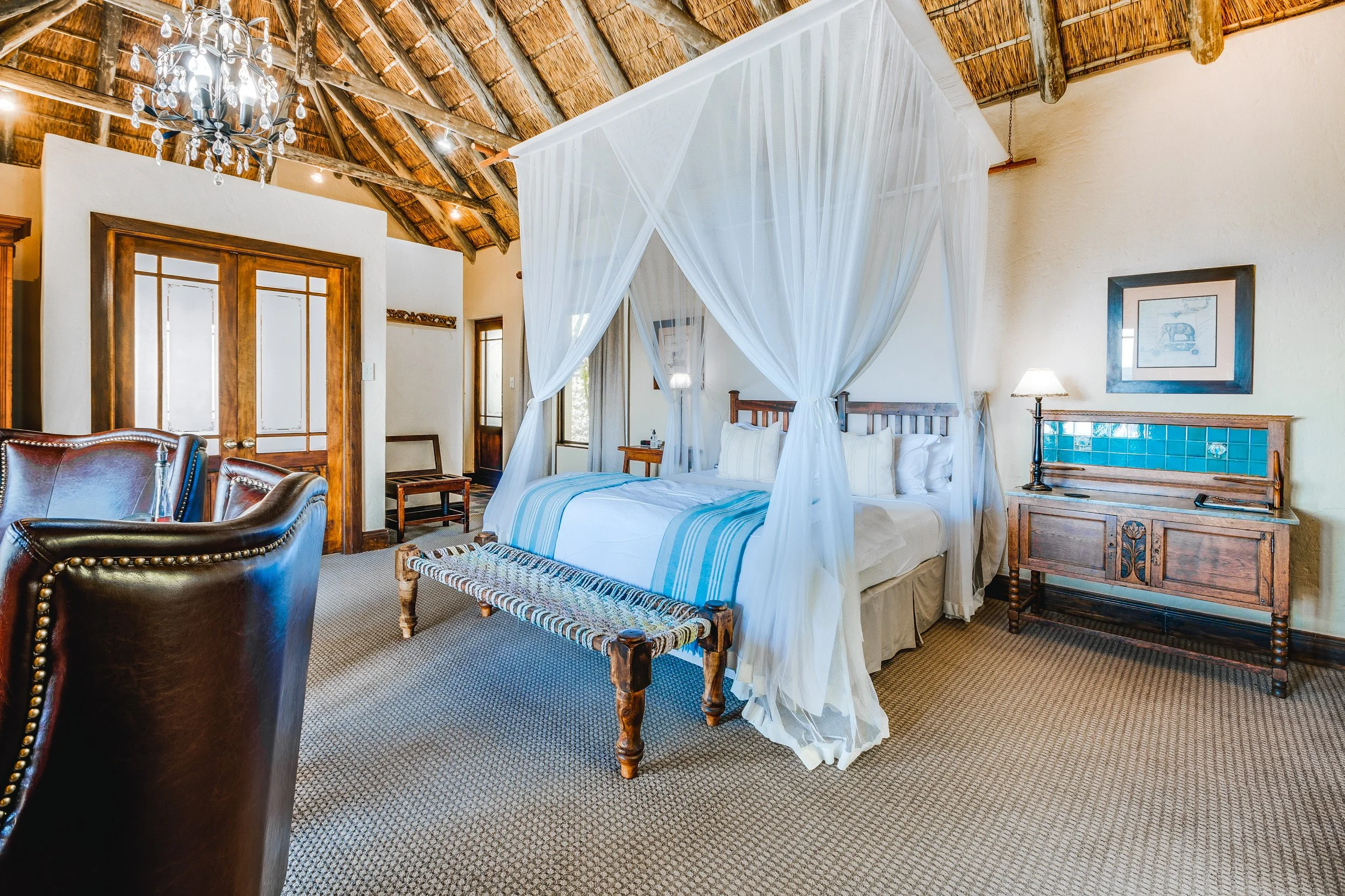 A bedroom with a sloped thatched roof, featuring a four-poster bed with white drapes and white bedding with blue stripes. There are two visible armchairs, a bench at the foot of the bed, a wooden dresser with blue tile accents, and framed artwork on the wall. A chandelier hangs from the ceiling.